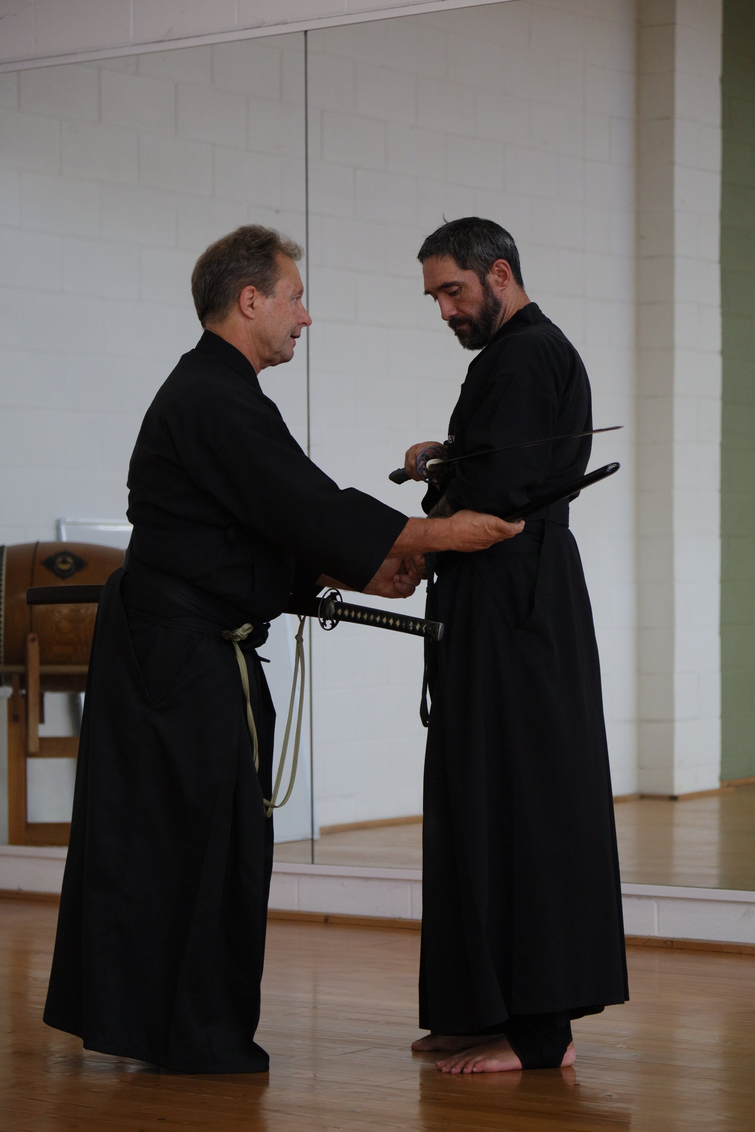 Two men in black martial arts uniforms, one holding a sword, standing indoors on a wooden floor, with one man barefoot and another with shoes off.