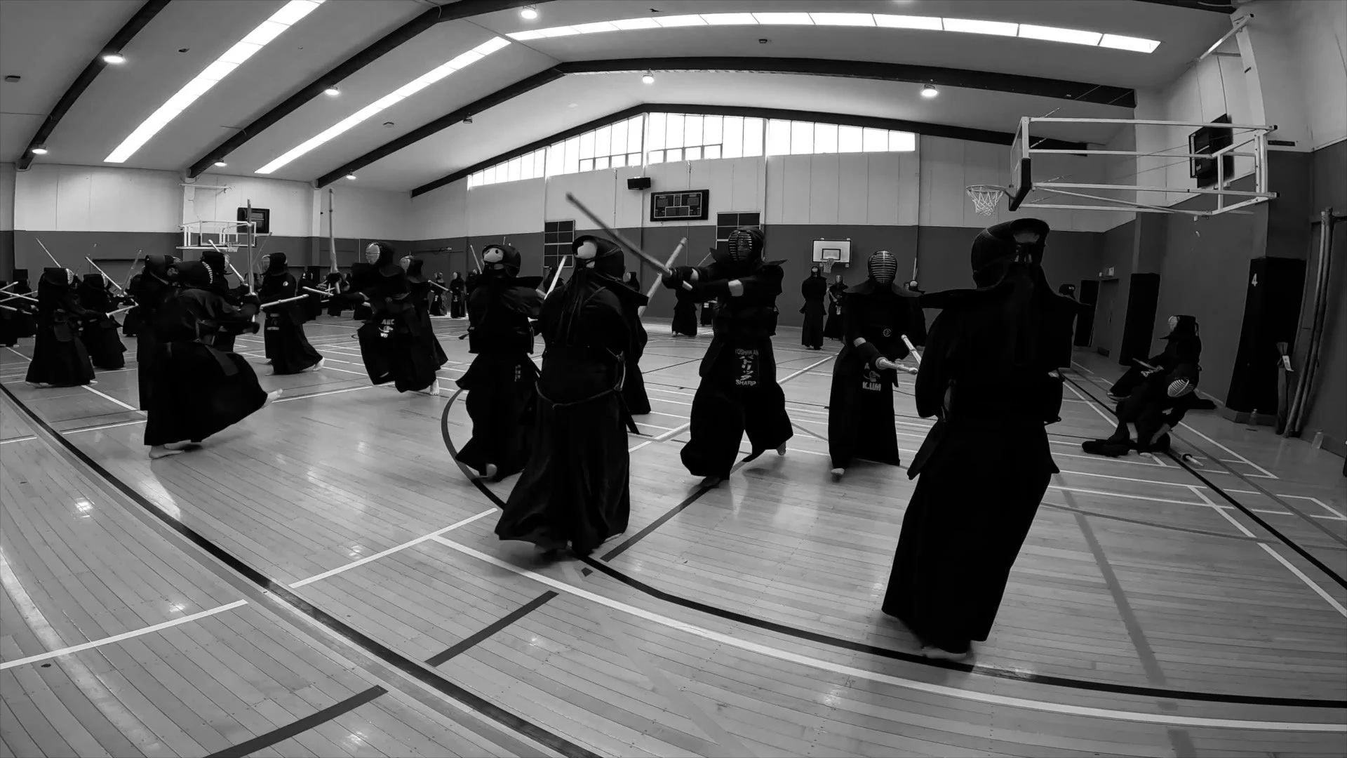 A large indoor gymnasium with a high ceiling and multiple windows, filled with people practicing Kendo, a Japanese martial art, dressed in traditional armor and uniforms.