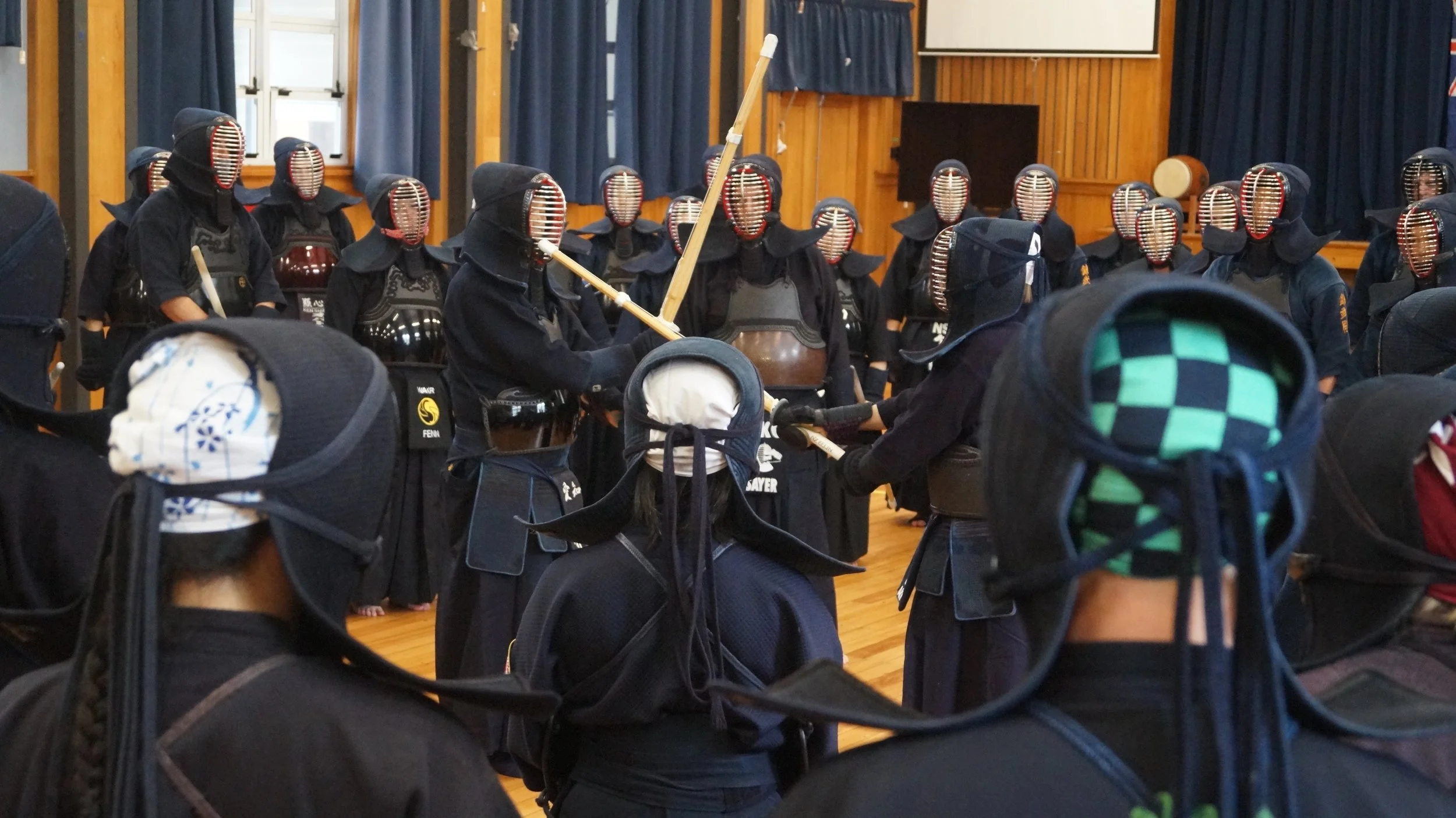 A group of people dressed in kendo gear, including helmets with face guards, gathered in a room with wooden walls and blue curtains, during a martial arts practice or event.