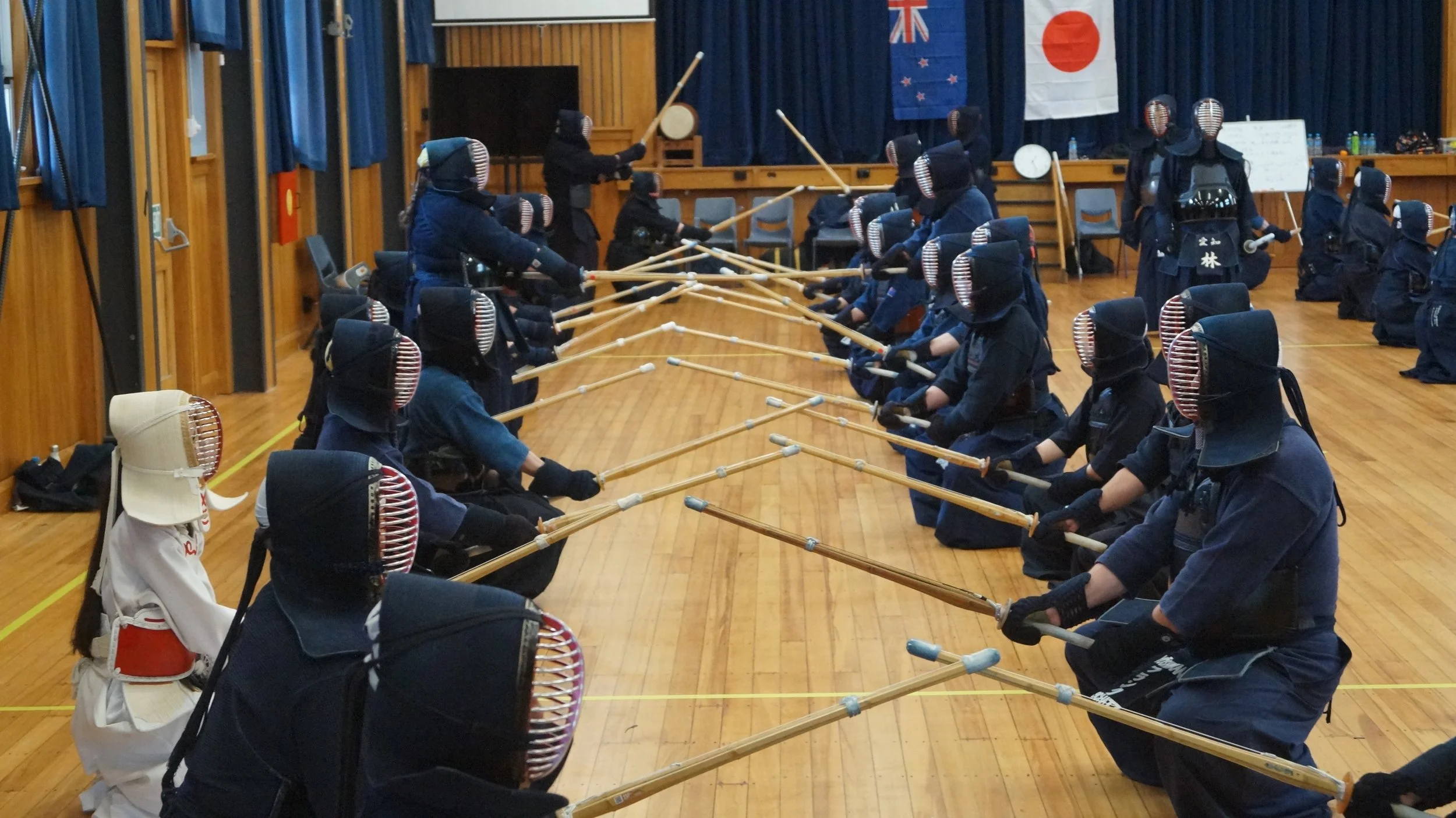 A group of people practicing Kendo in a gymnasium with Japanese flags on the background. They are dressed in traditional Kendo armor and holding bamboo swords, engaging in a training exercise.