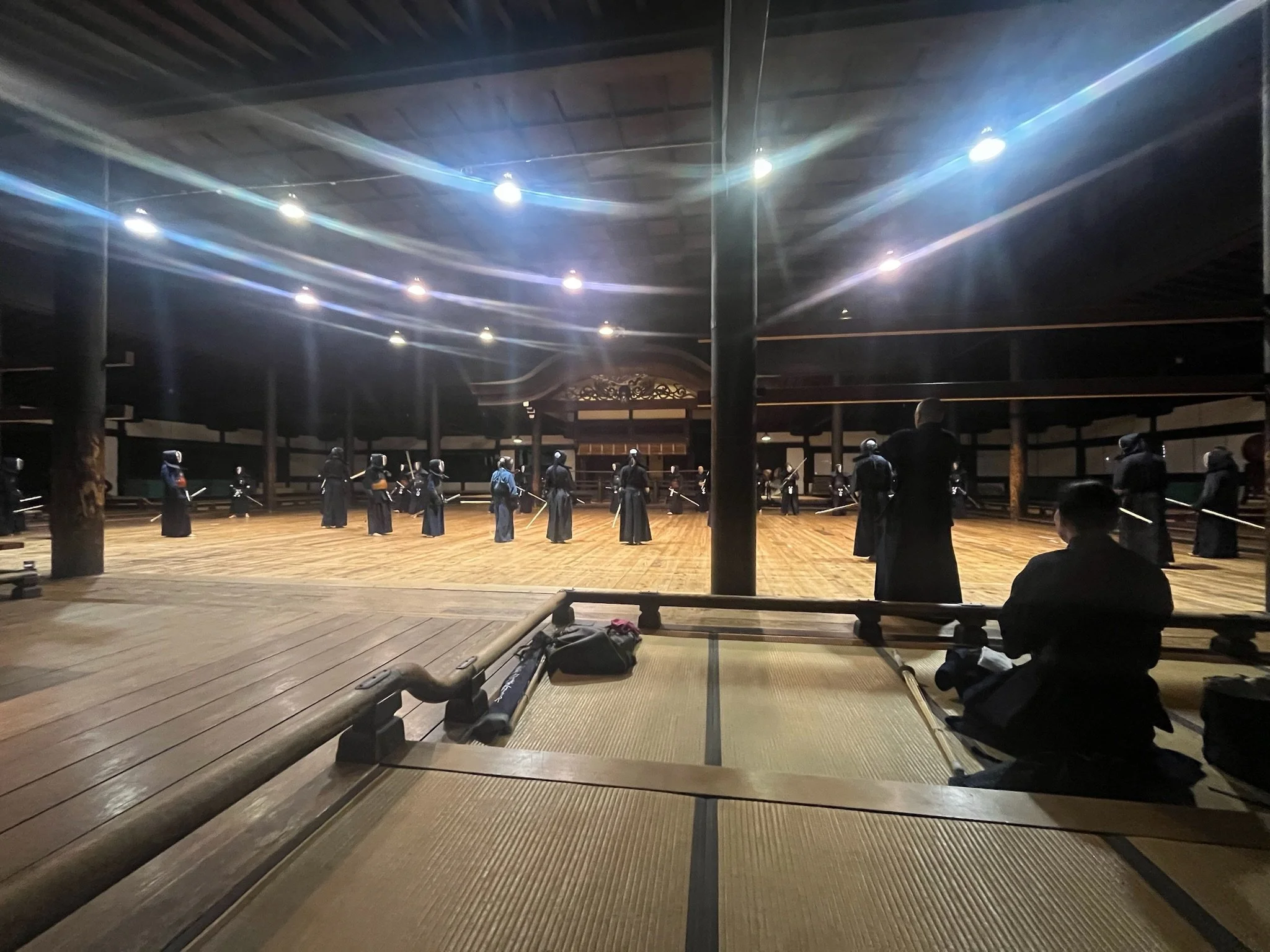 A dojo or martial arts training hall with students dressed in traditional martial arts uniforms (karate or kendo) practicing or preparing for training, illuminated by ceiling lights.