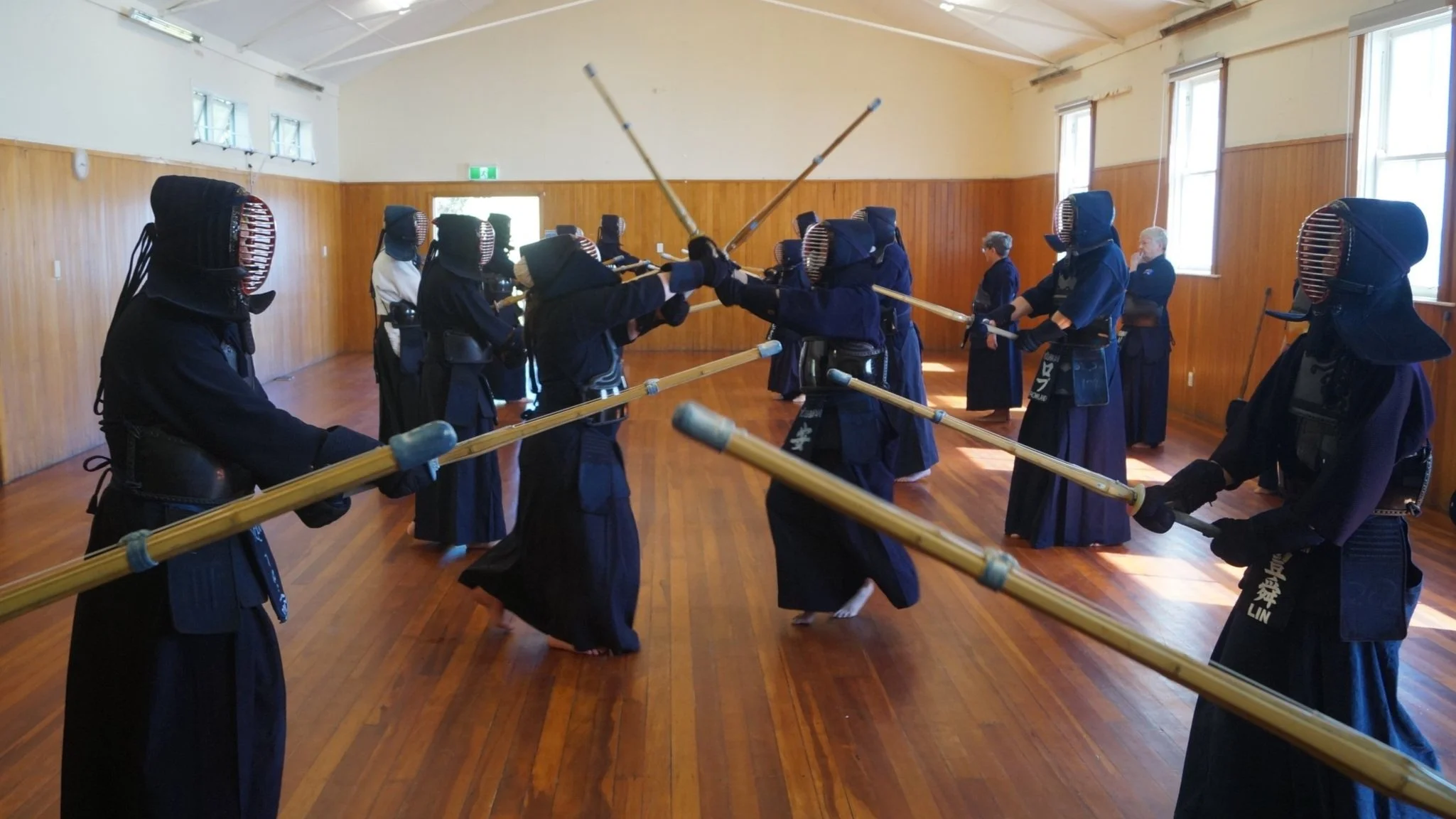 A group of people practicing Kendo, a traditional Japanese martial art, in a wooden-floored dojo. They are wearing protective gear and holding bamboo shinai swords.