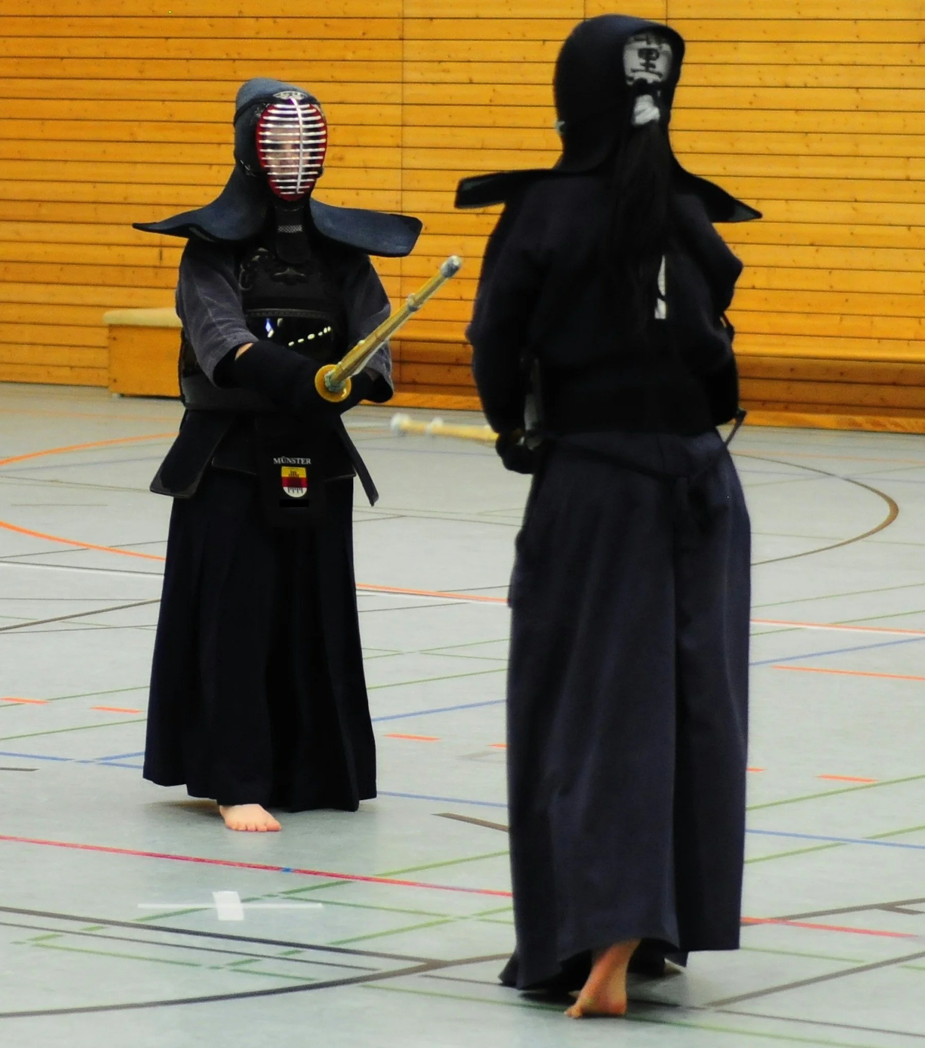 Two individuals practicing kendo or a similar martial art in a gymnasium with wooden wall panels. 