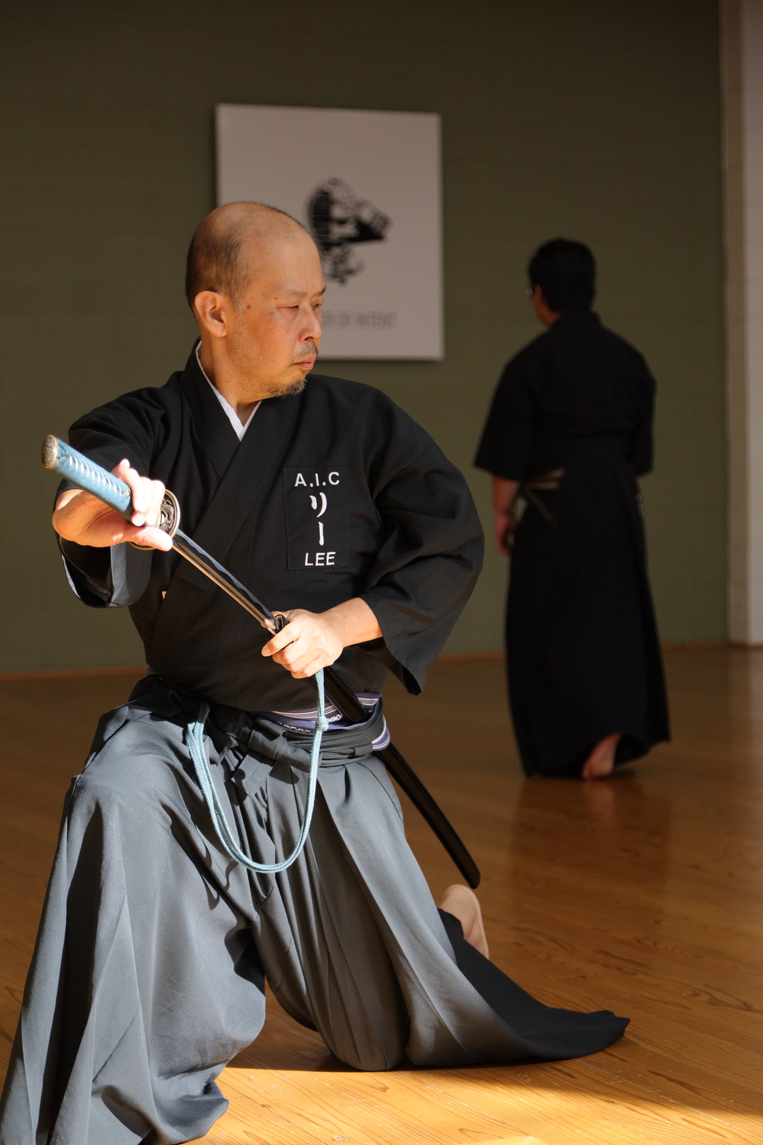 A man in traditional martial arts attire kneeling on the wooden floor, holding a katana sword, in a martial arts dojo with another person in the background.