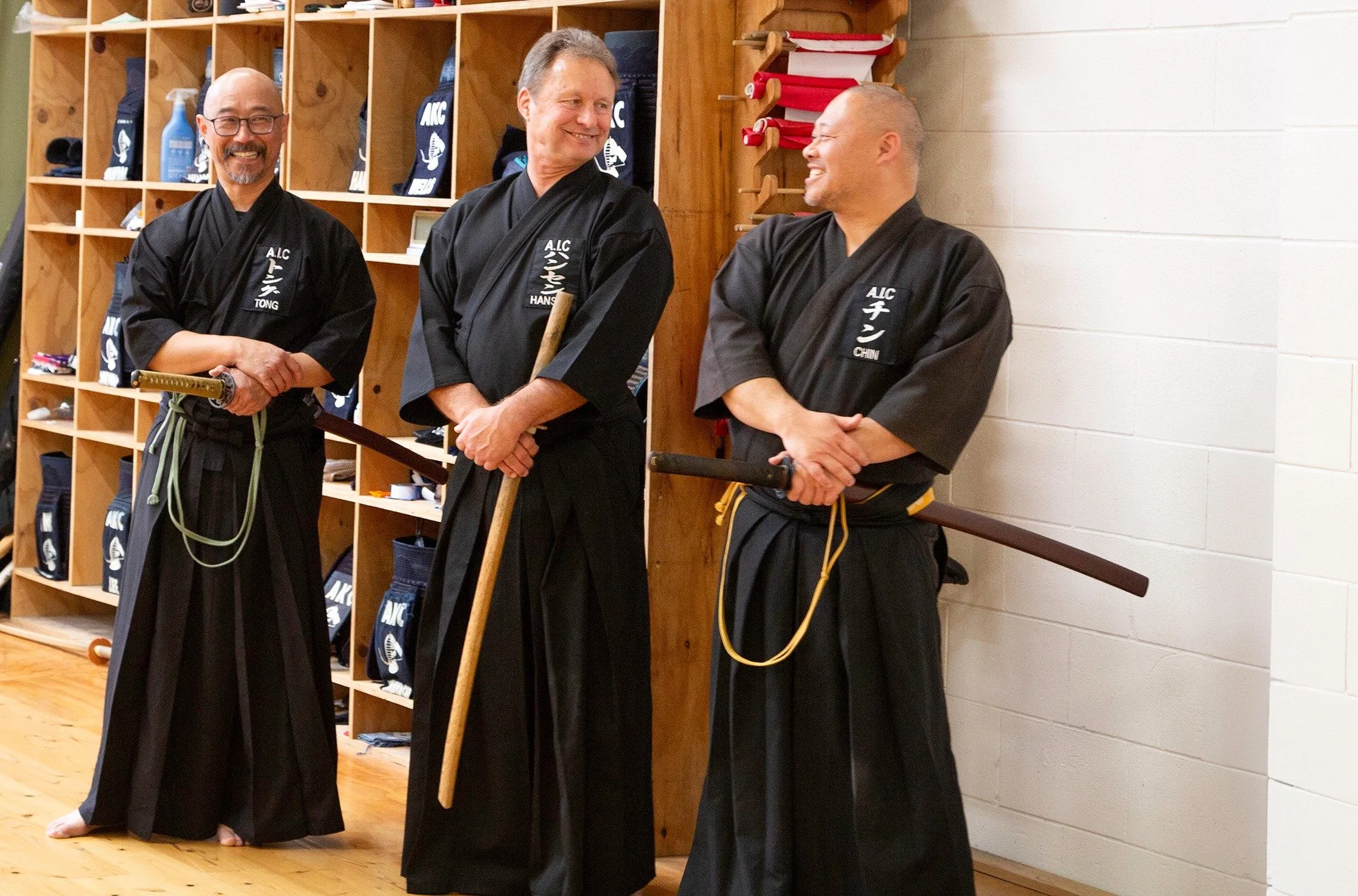 Three men dressed in black martial arts uniforms standing together in a dojo, smiling and laughing, with shelves of gear and white cinder block walls in the background.