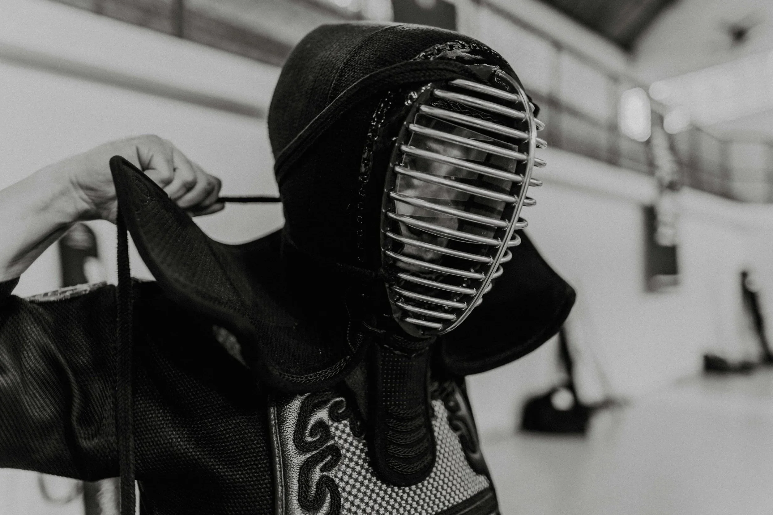 A person wearing a kendo helmet and protective gear, holding a shinai, a bamboo sword used in kendo, in an indoor martial arts training space.