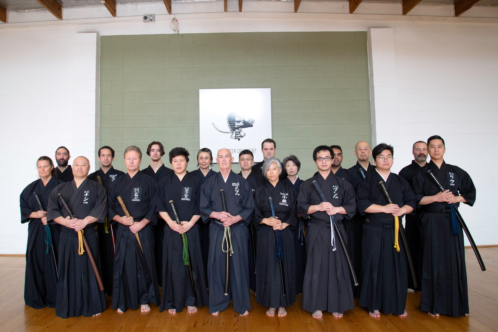 Group of martial artists in black uniforms standing in a gym, holding weapons, facing forward, with a white wall and green accent wall behind them.