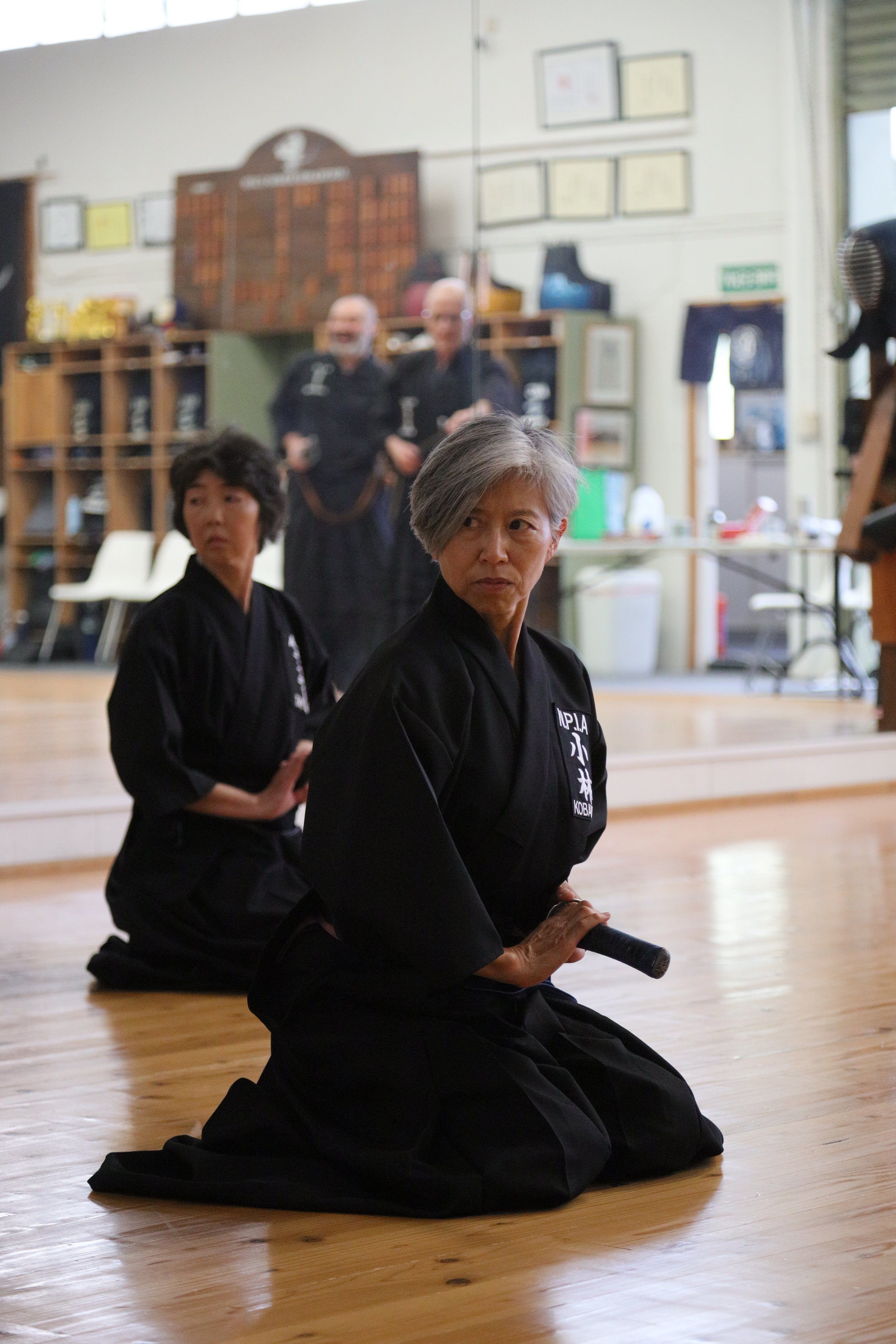 Three women wearing traditional black martial arts uniforms practicing martial arts in a dojo, with two women in the background and one in the foreground holding a katana, sitting on their knees on a wooden floor.