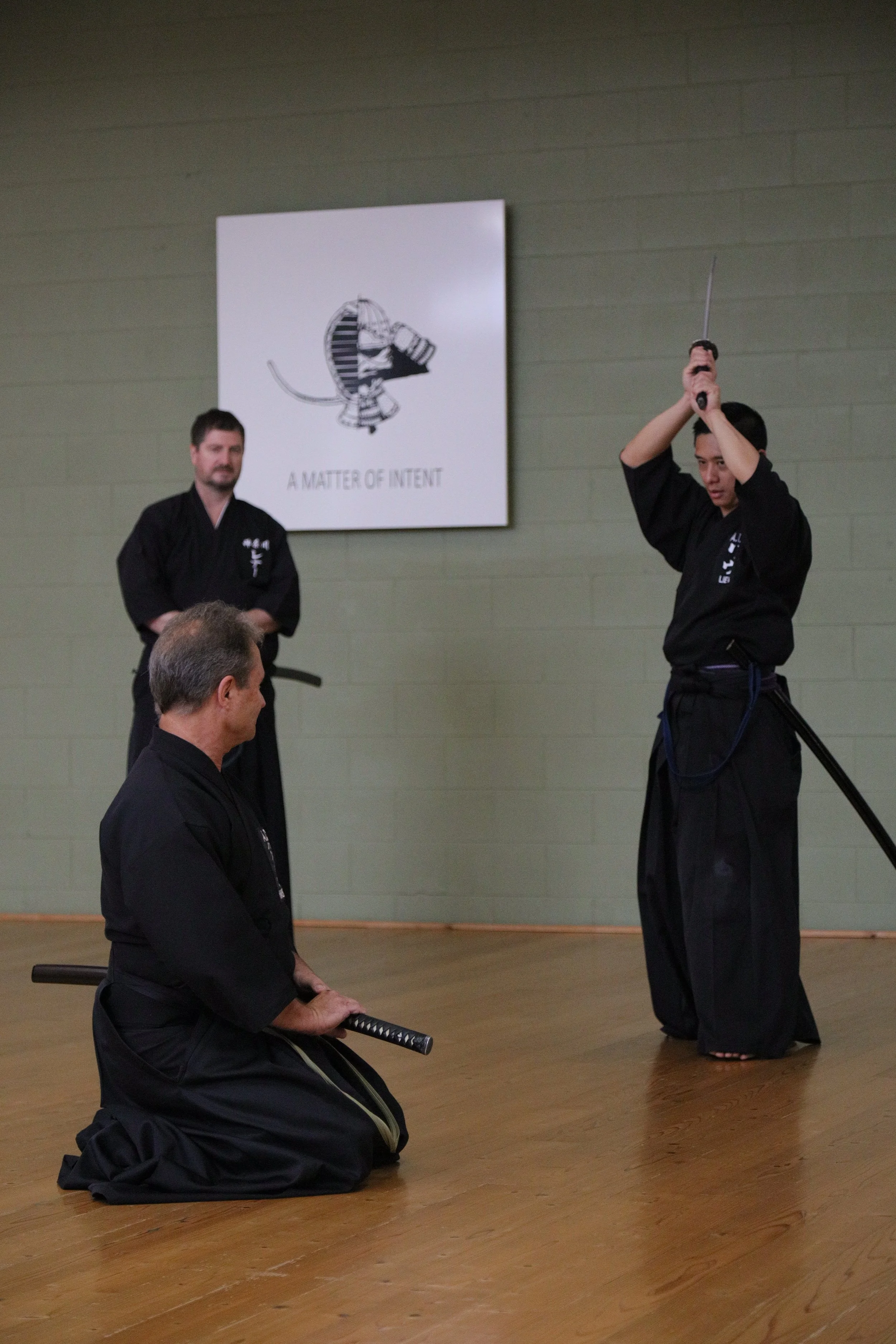 Two martial artists in traditional black uniforms practicing Kendo, with one kneeling and holding a sword, and the other standing and preparing to strike, in a dojo with a wooden floor and a poster on the wall reading "A Matter of Intent."