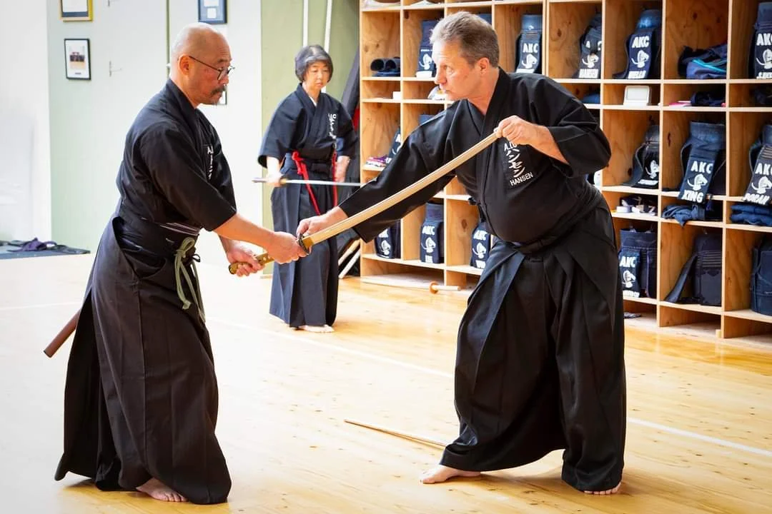 Two practitioners practicing Iaido in a dojo, with a third person observing in the background.