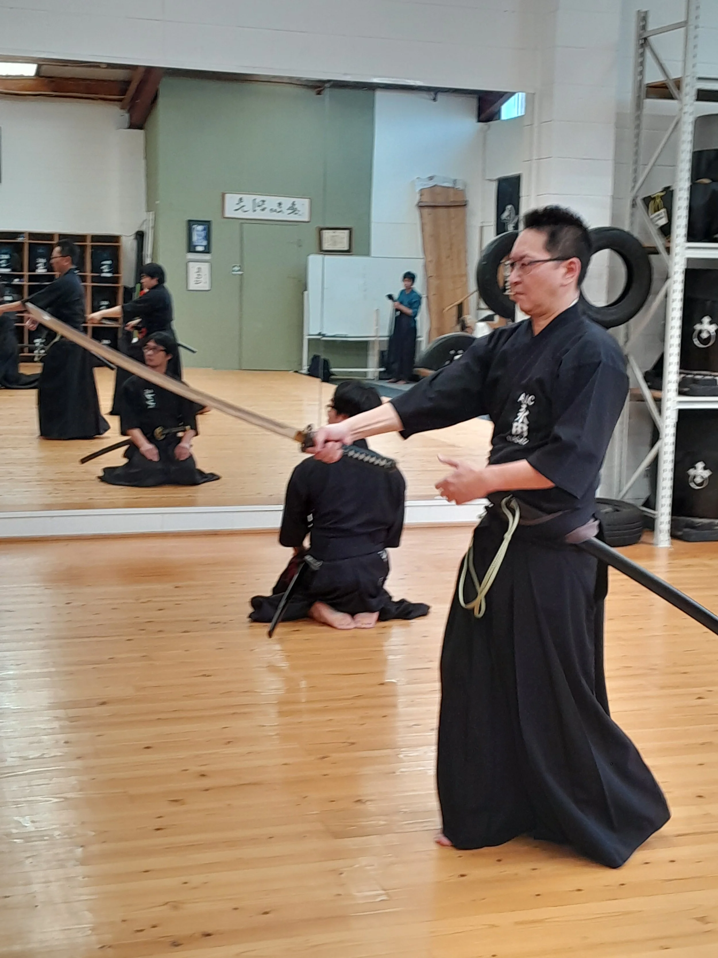Martial arts students practicing in a dojo, some kneeling and some standing, with one man in the foreground wearing traditional black gi uniform holding a katana sword.