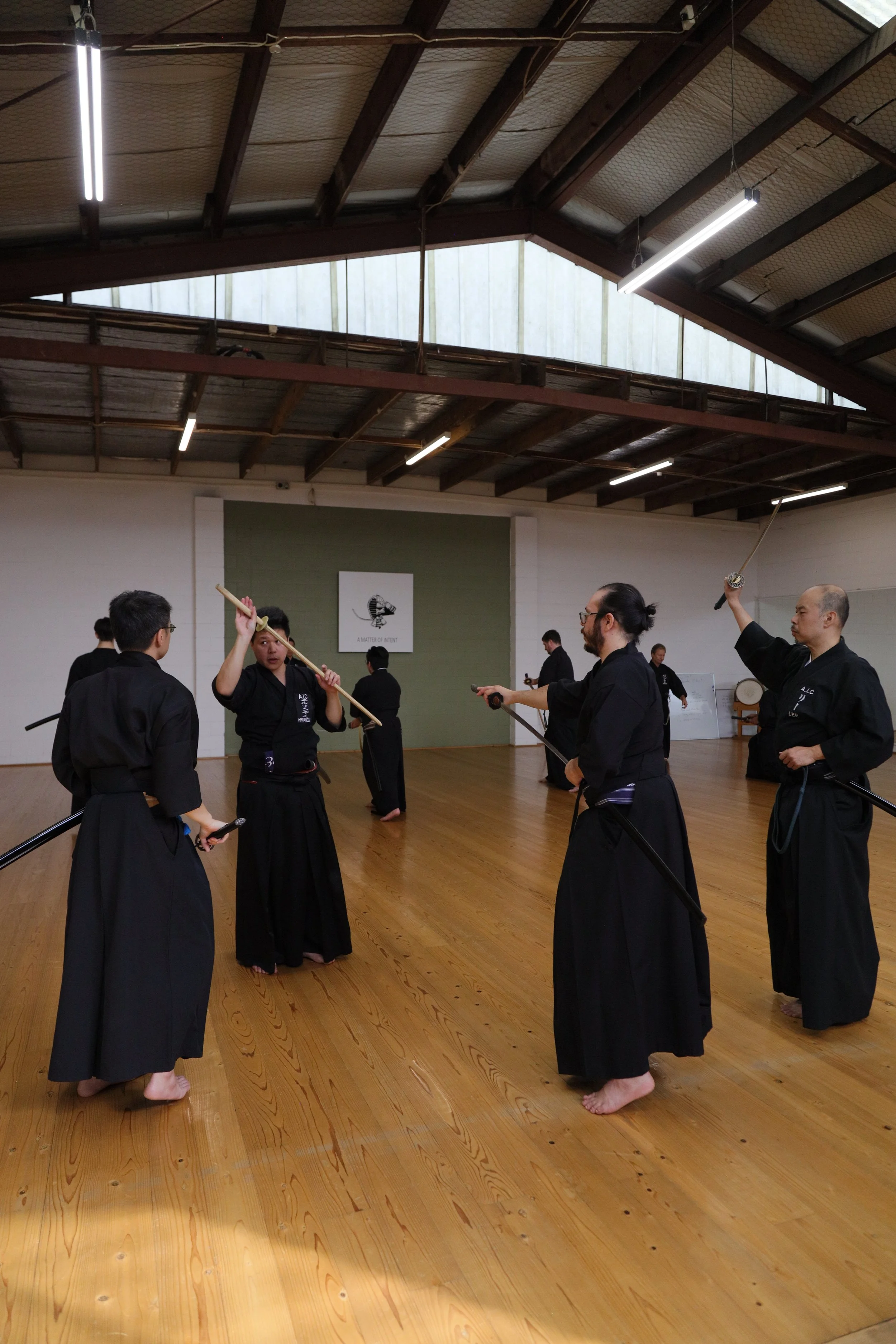 A group of martial artists practicing with swords in a dojo, wearing traditional black uniforms, with some holding swords and others observing.