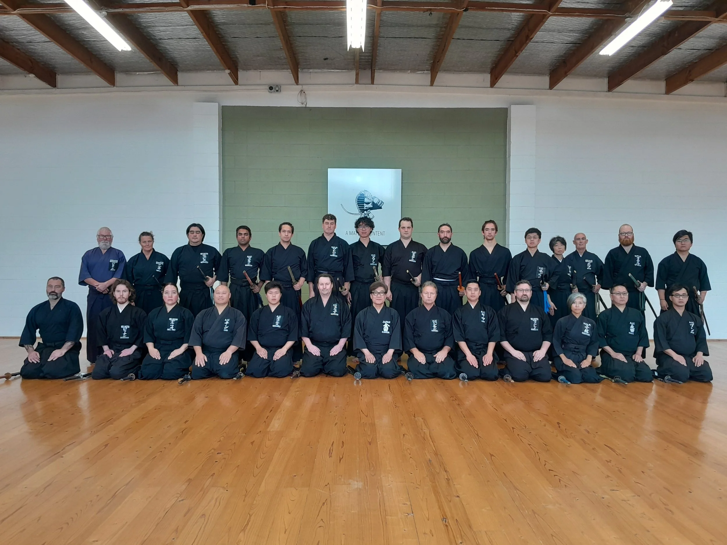 Group photo of martial arts students and instructors in a dojo, dressed in black uniforms, standing and kneeling in front of a green wall with a martial arts emblem and text.