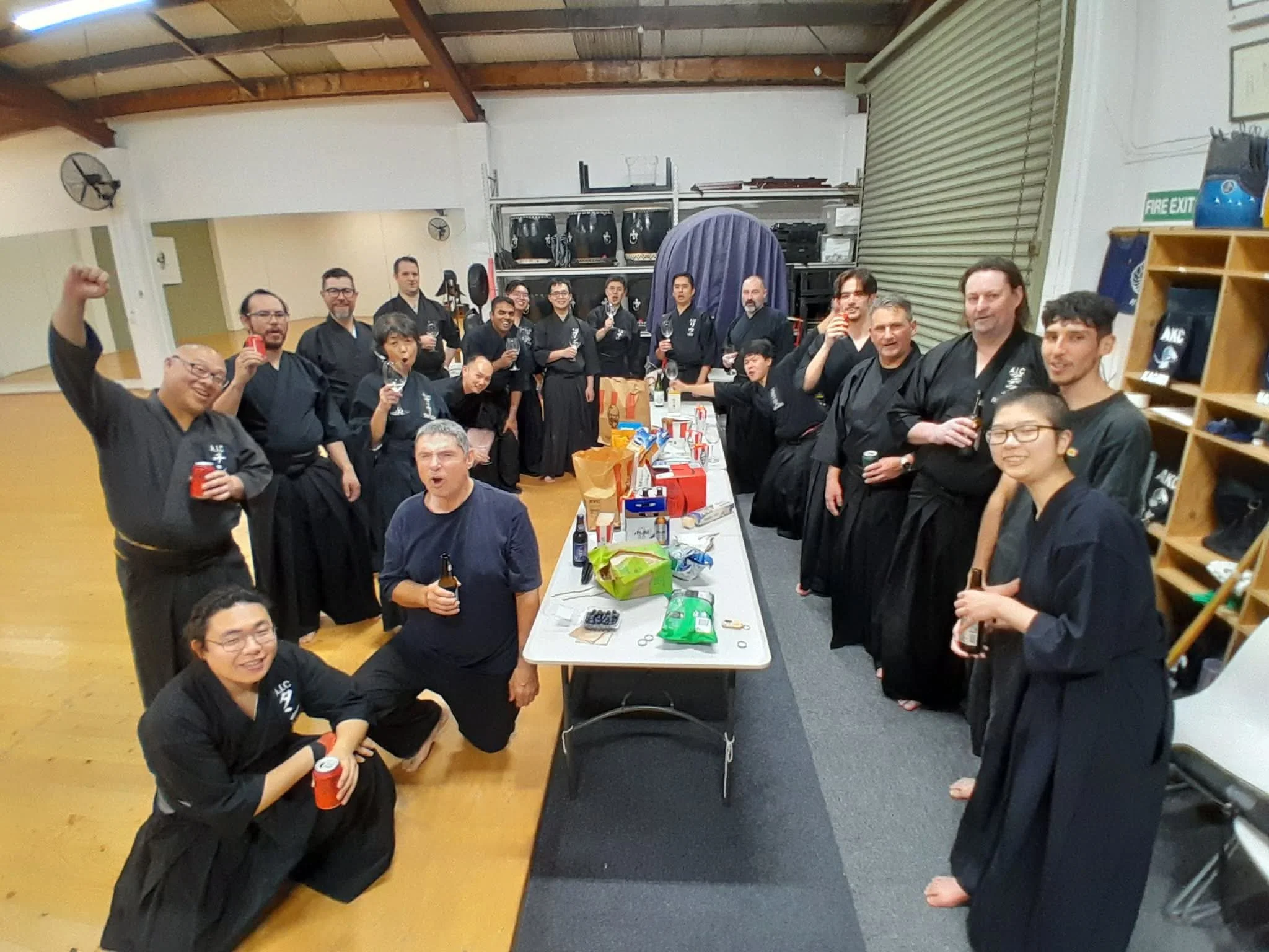 Group of people in black martial arts uniforms gathered around a table with snacks and drinks in a warehouse or industrial space, celebrating.