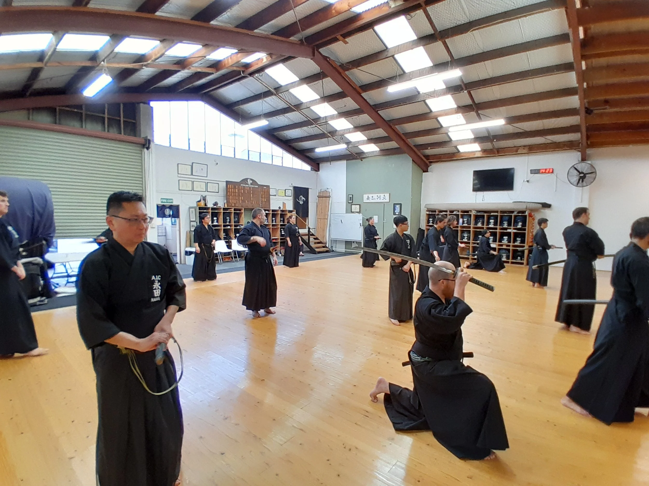 Martial arts class in a dojo with students in black uniforms practicing Iaido, some students kneeling and others standing.