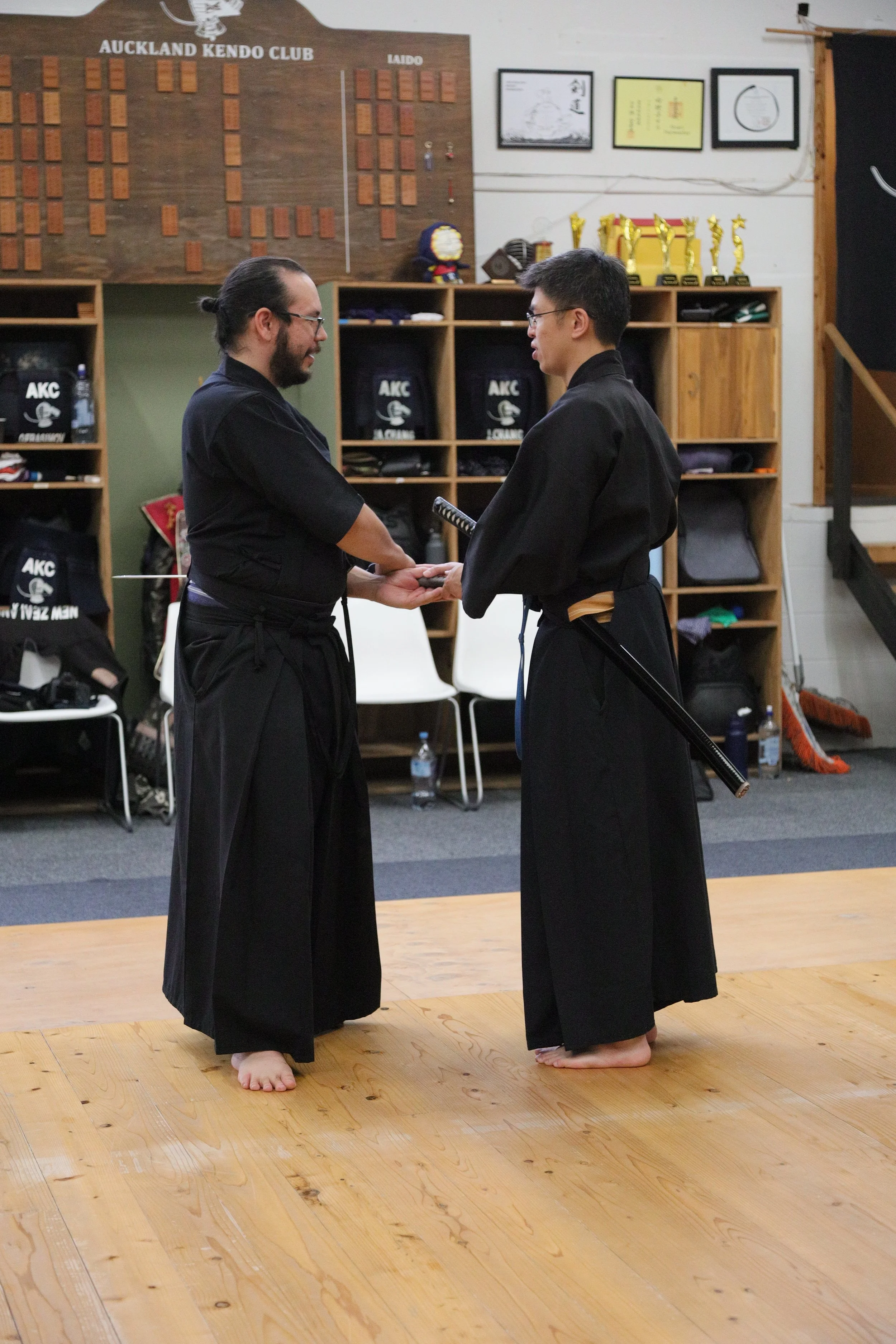 Two men practicing martial arts, wearing traditional black uniforms, standing barefoot on a wooden floor, facing each other and holding hands in a dojo with awards, trophies, and framed pictures on the walls.