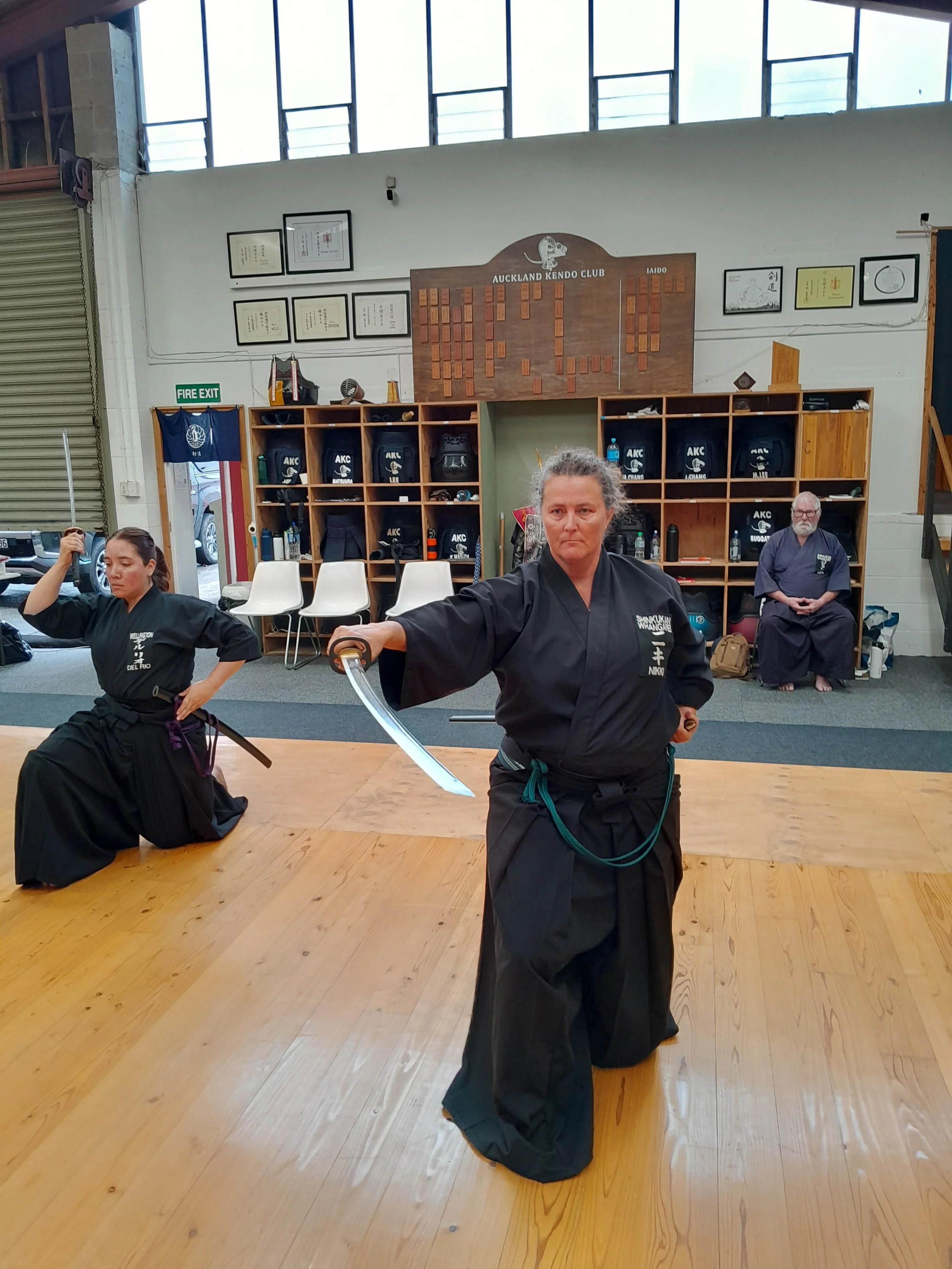 Martial arts practitioners practicing in a dojo, with a woman in the foreground holding a sword and others in martial arts uniforms.