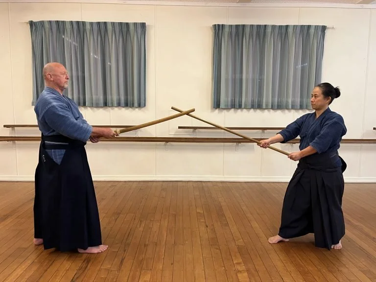 Two martial artists practicing jodo with wooden weapons in a dojo.