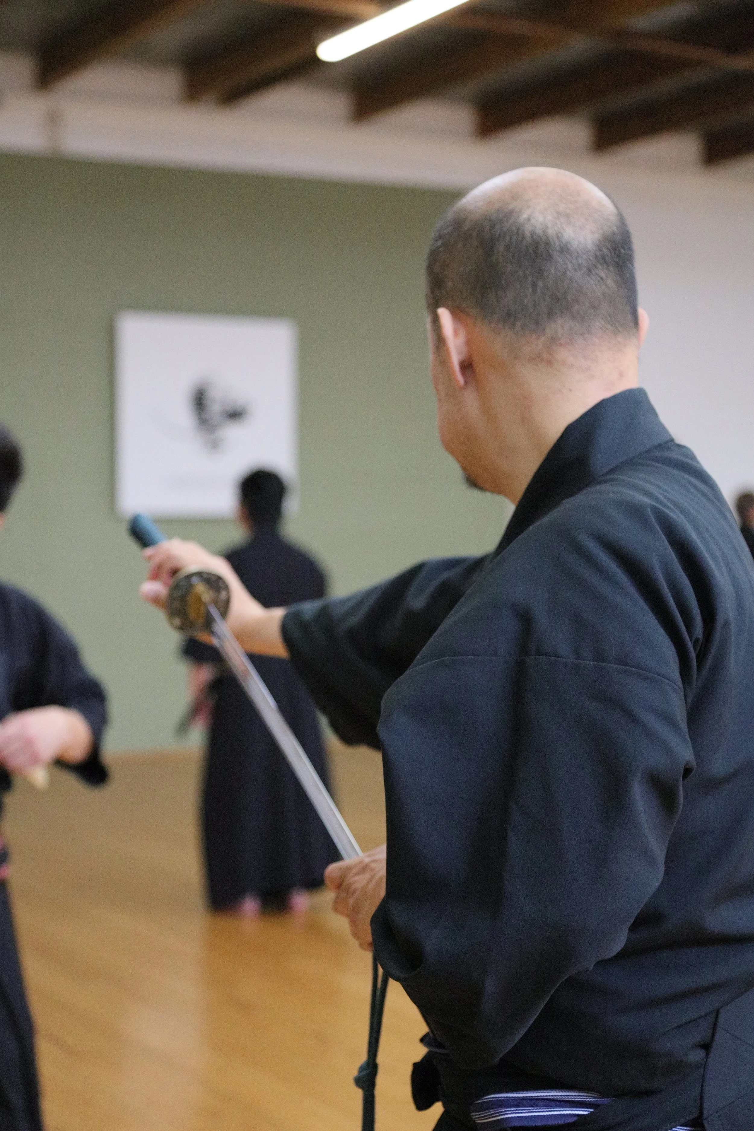A man in traditional martial arts attire is holding a sword during a martial arts demonstration or class. He is facing away from the camera, with a group of people in the background.
