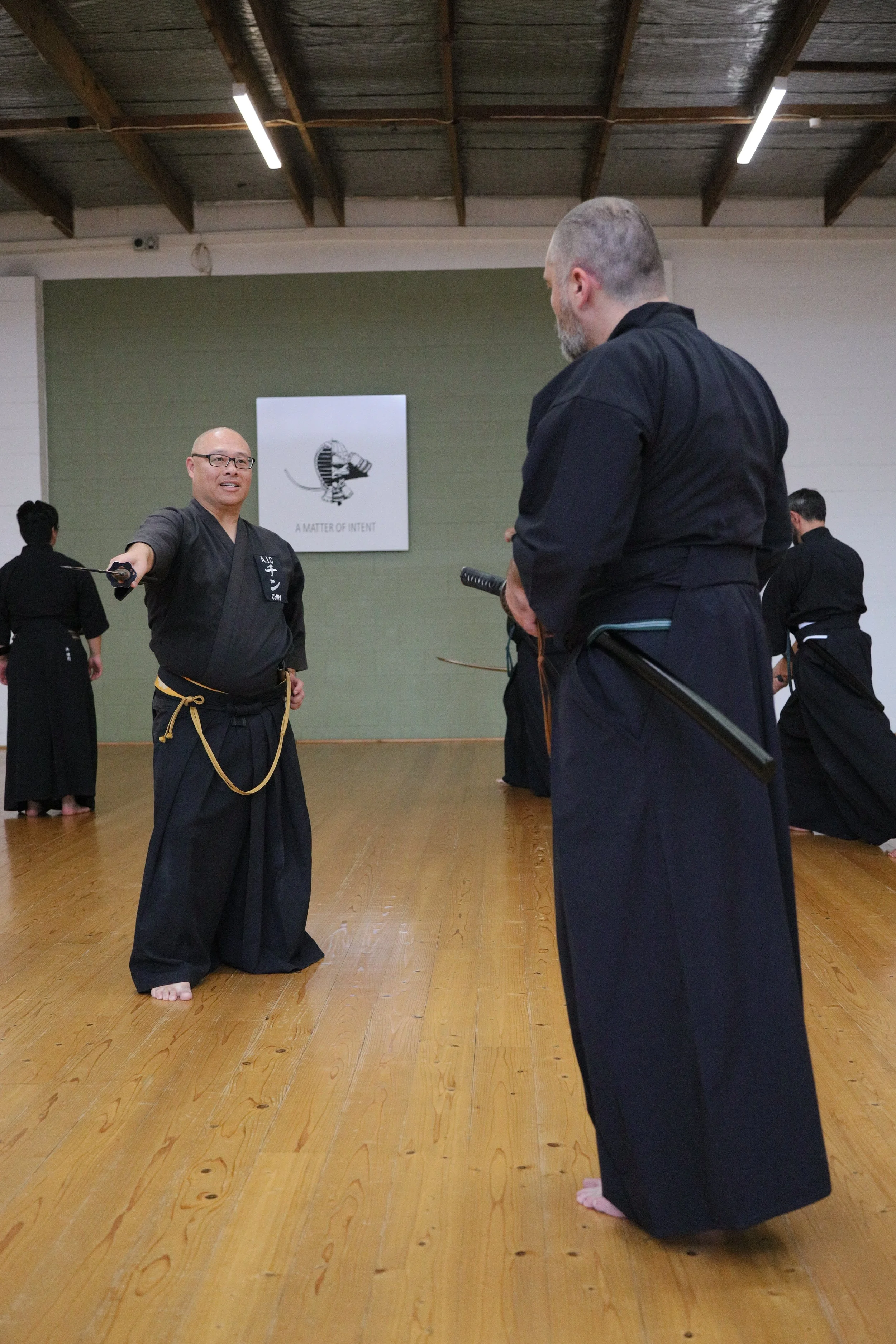 Two men practicing martial arts in a dojo. One man, with glasses, is holding a katana and pointing it towards the other man, who has a beard. Both are wearing black martial arts uniforms. Other individuals in black uniforms are visible in the backgro