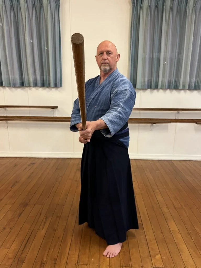 A man in traditional martial arts attire holding a wooden staff in a dojo with wooden floor and ballet barres along the wall.