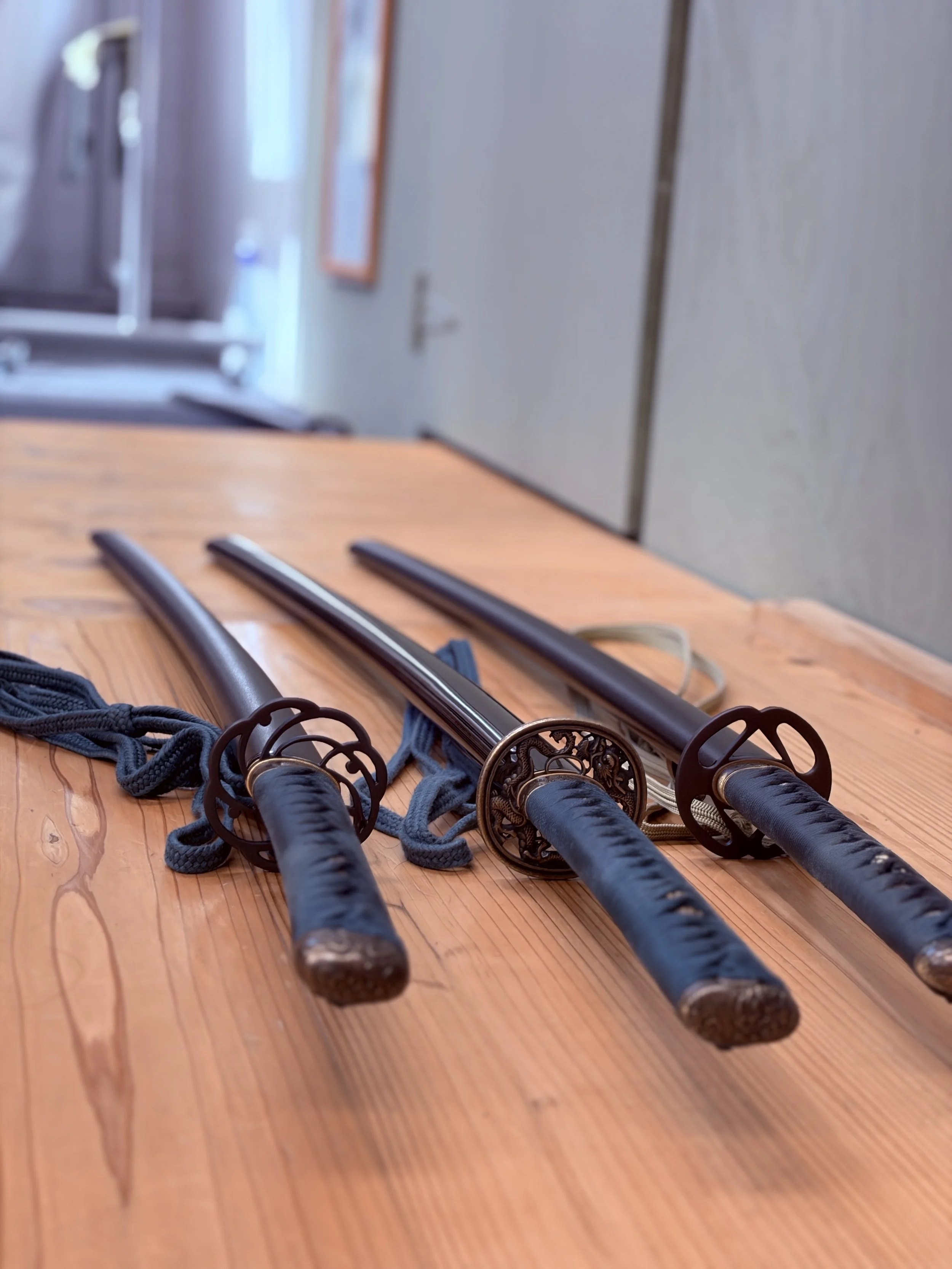 Three Japanese katanas with black scabbards and ornate hand guards resting on a wooden table.