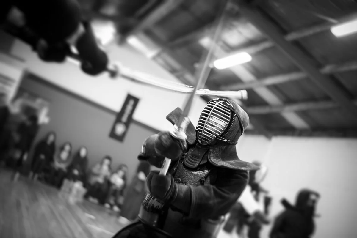 A person dressed in fencing gear, including a helmet and padded protective clothing, is holding a fencing sword in a fencing stance. The background shows several spectators watching in an indoor gym or sports hall.