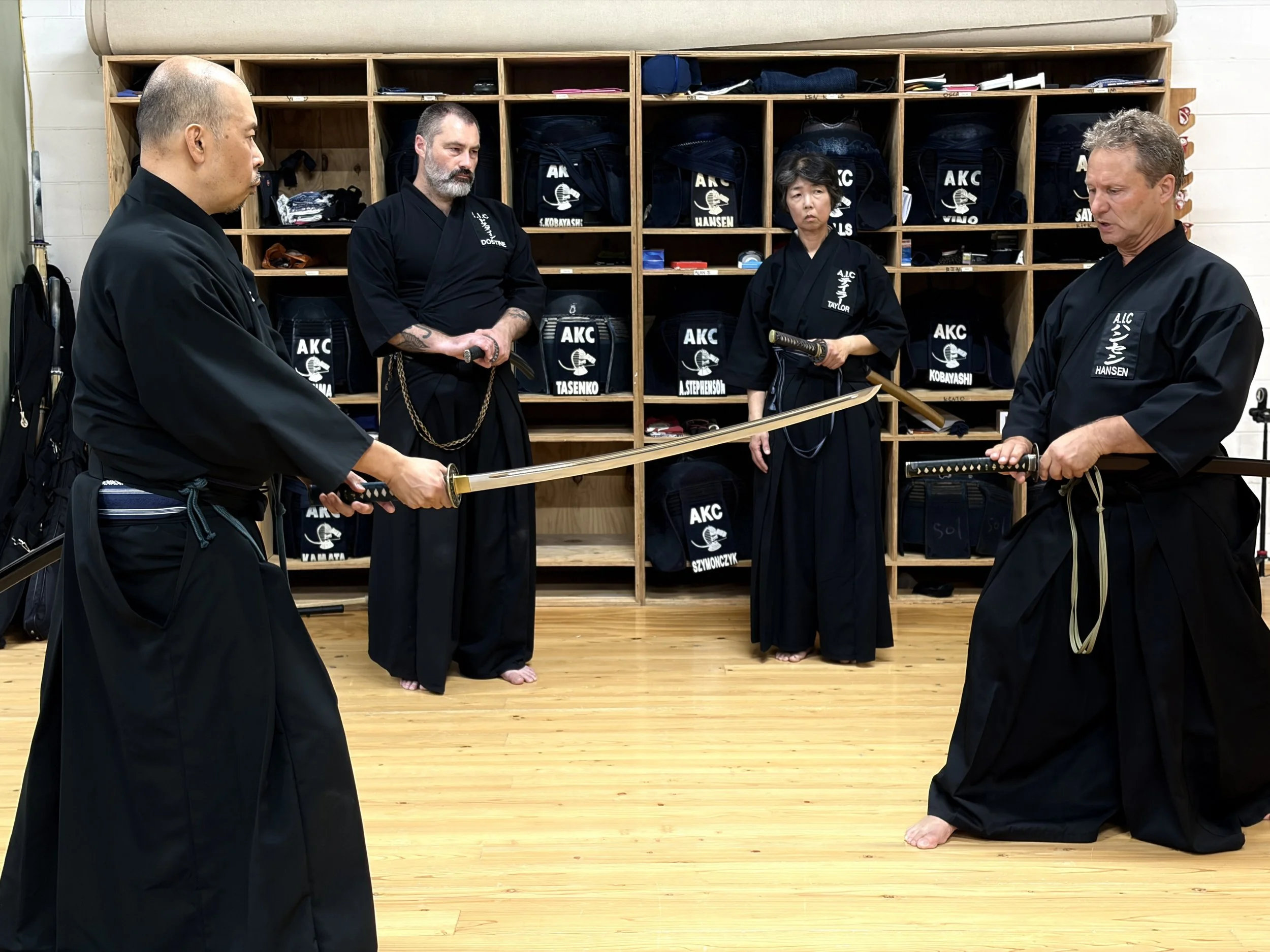 Five martial artists in traditional black uniforms with belts and swords are practicing in a dojo, standing on a polished wooden floor in front of wooden shelves filled with bags and gear.