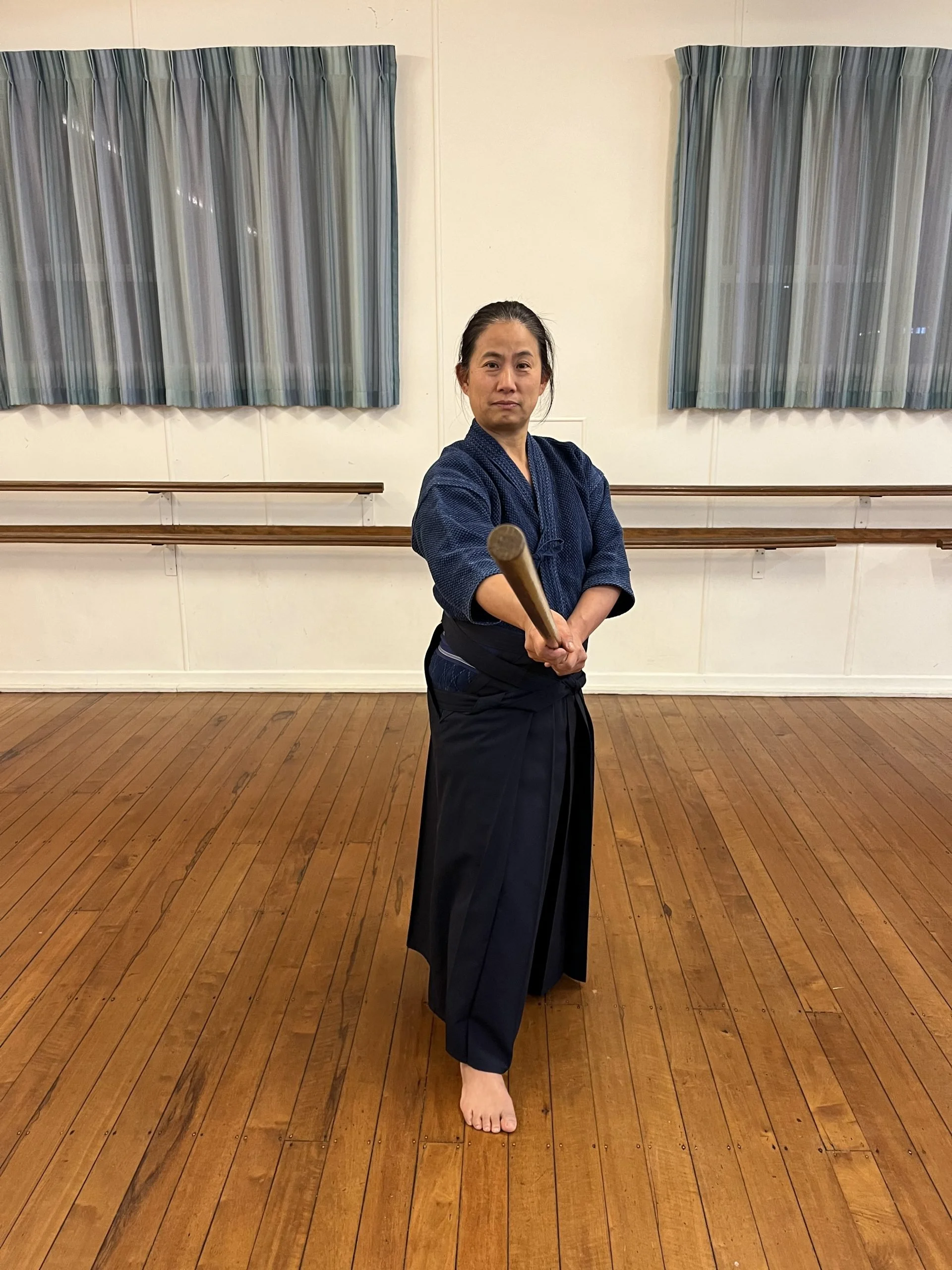 A person dressed in traditional martial arts attire, holding a wooden staff in a martial arts pose in a dojo.