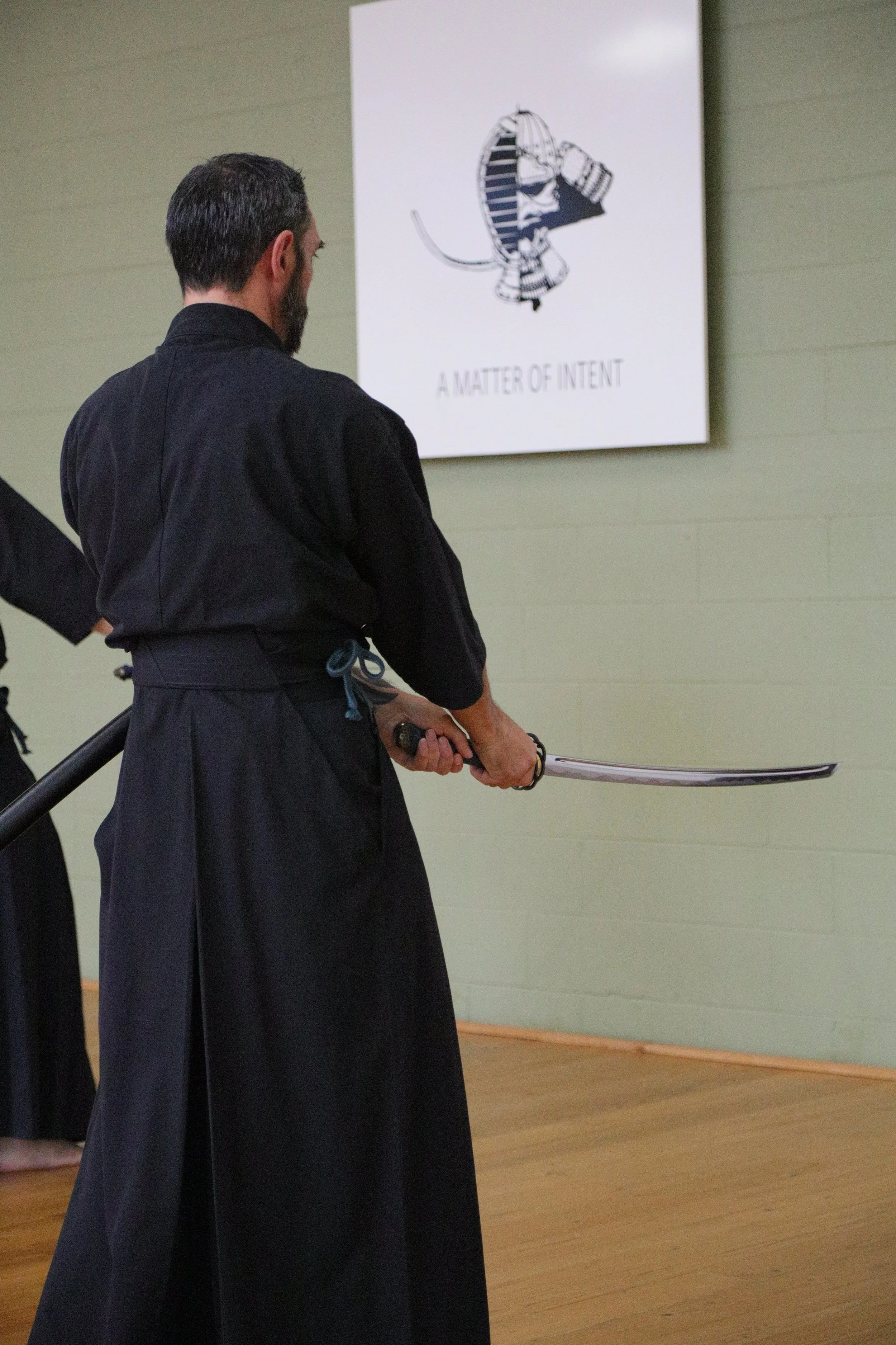 A person dressed in traditional black martial arts gi holding a sword, standing indoors in front of a poster that features an illustration of a mouse dressed in a similar martial arts outfit, with the text "A MATTER OF INTENT". The setting appears to