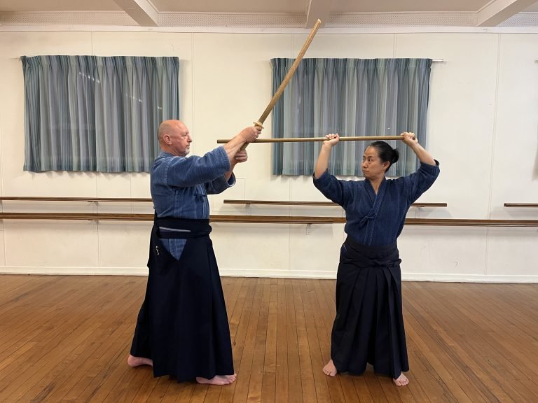 Two martial artists practicing with wooden staffs in a dojo, one older man and one younger woman, both wearing traditional kimono-style uniforms and standing on a wooden floor, with a mirror and curtains on the wall behind them.