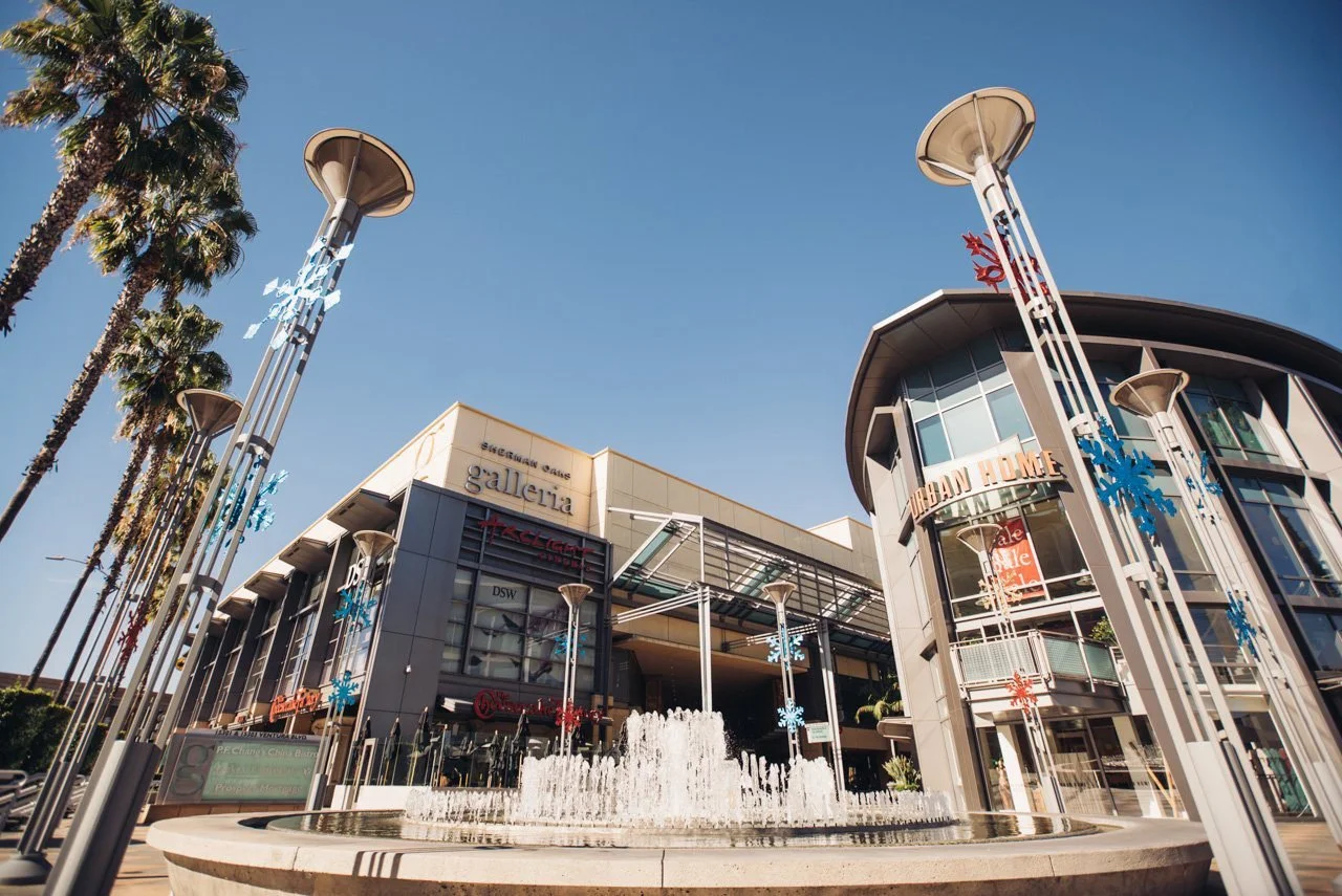 Sherman Oaks shopping mall with a water fountain in front, surrounded by tall palm trees. The mall features multiple levels with modern architecture, including glass windows, and holiday decorations such as snowflakes and red-and-blue ornaments.