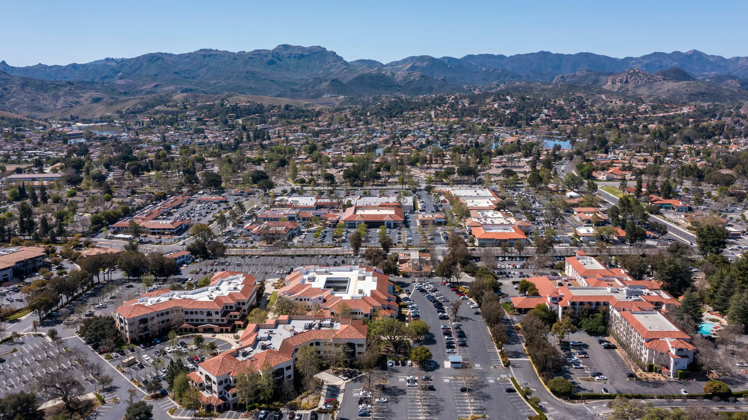 Thousand Oaks, CA aerial view of a suburban shopping area with multiple buildings, parking lots, and tree-lined streets, surrounded by mountains in the background.