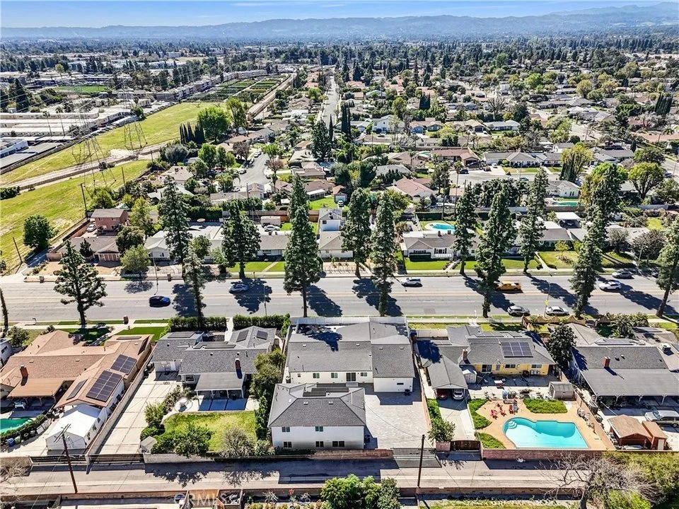 Aerial view of a residential neighborhood with single-family homes, a street with cars, tall trees, and backyard pools, with mountains in the background and a clear sky.