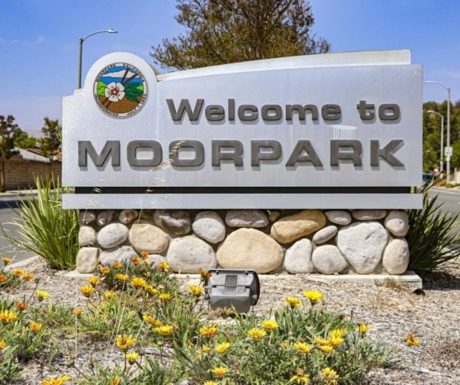 A welcome sign for Moorpark with a landscape emblem at the top, stone base, surrounded by flowers and plants, next to a street with streetlights and houses in the background, serving Moorpark