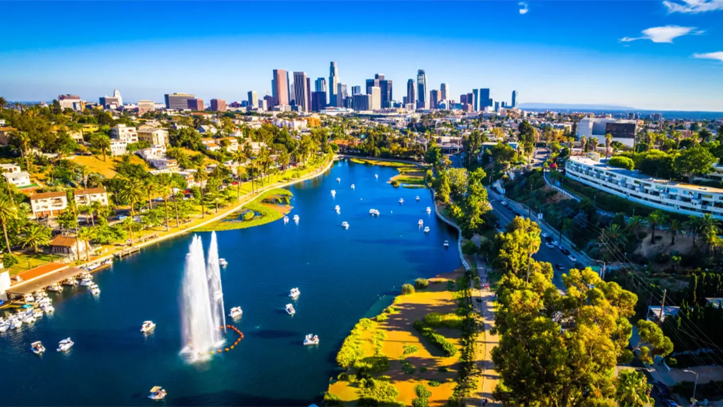 Aerial view of a Los Angeles skyline with tall skyscrapers, a large pond with boats and a fountain, surrounded by lush green trees, residential areas, and a highway.