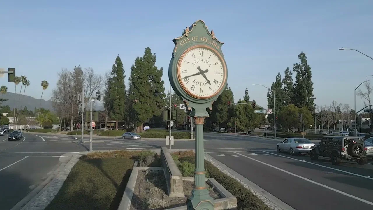 Arcadia, CA, vintage-style clock on a post , with the city name 'Arcadia' on the clock face, situated at an intersection with cars and traffic lights, surrounded by trees and buildings in the background.