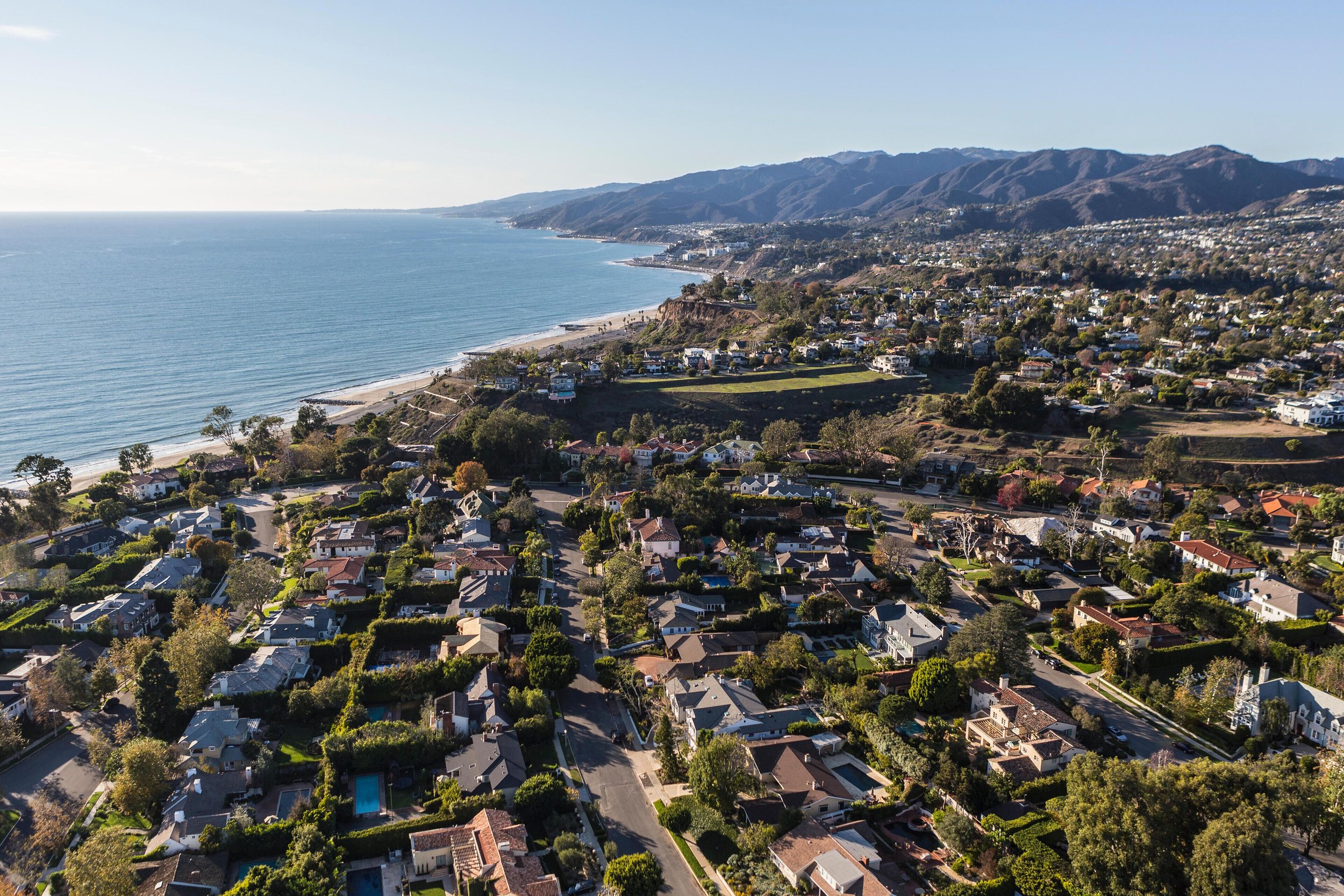Pacific Palisades aerial view of a coastal residential neighborhood with houses, trees, and streets, overlooking the ocean with sandy beach and distant mountains under a clear blue sky.