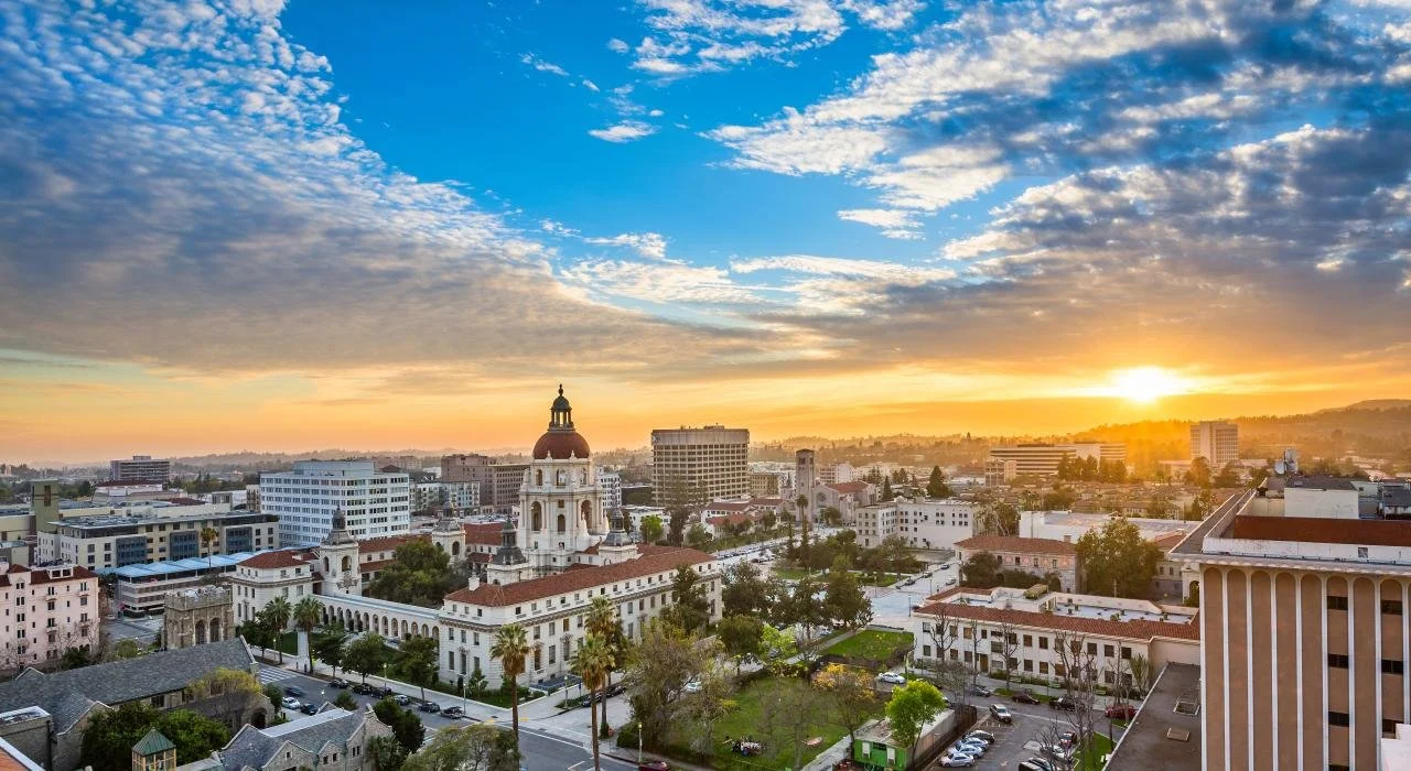 A cityscape at sunrise with historic Pasadena, CA buildings featuring a dome and bell tower, surrounded by modern buildings, trees, and a partly cloudy sky.