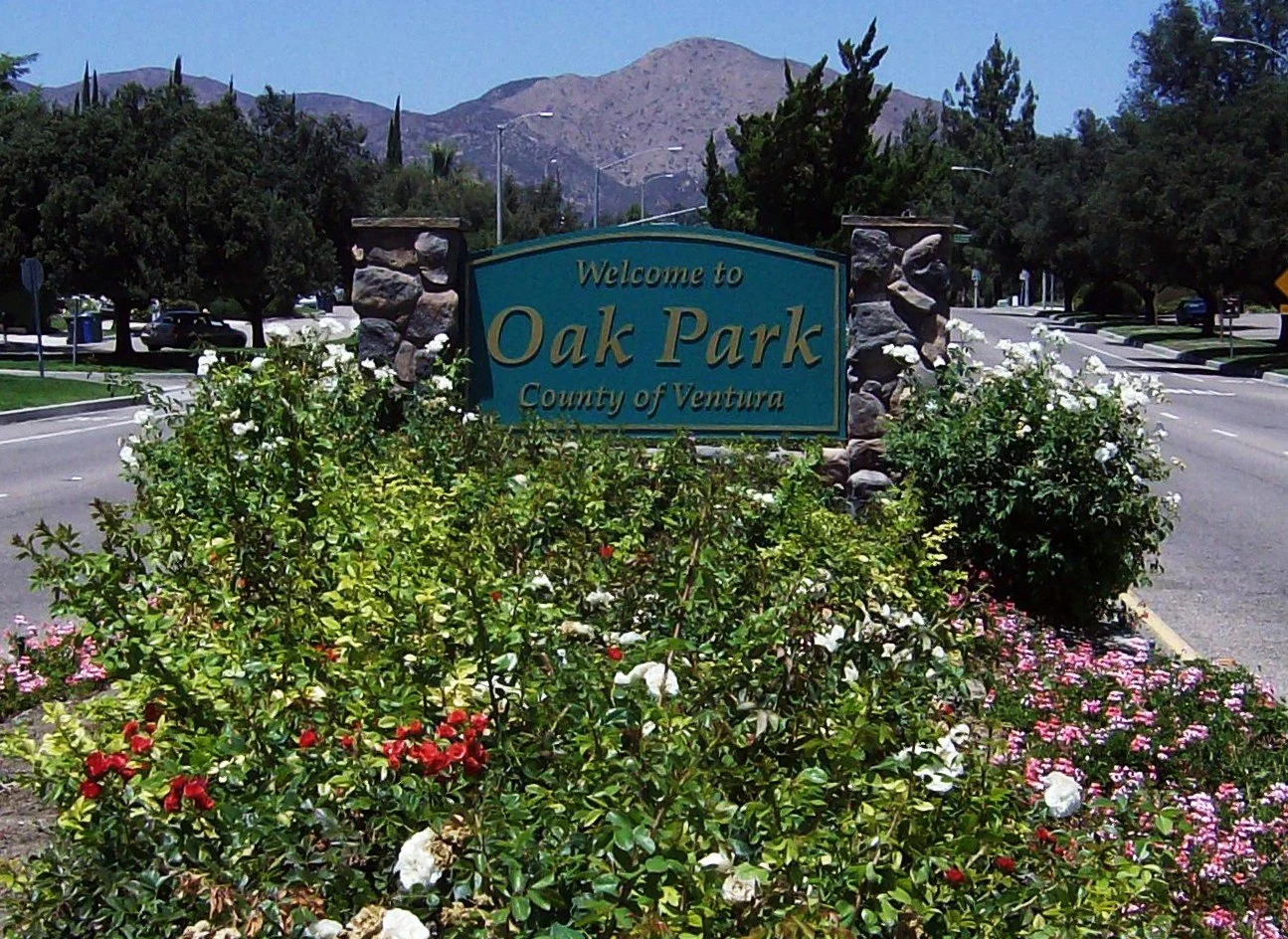Welcome sign for Oak Park in California, surrounded by colorful flowers and greenery. Mountains and trees are visible in the background.