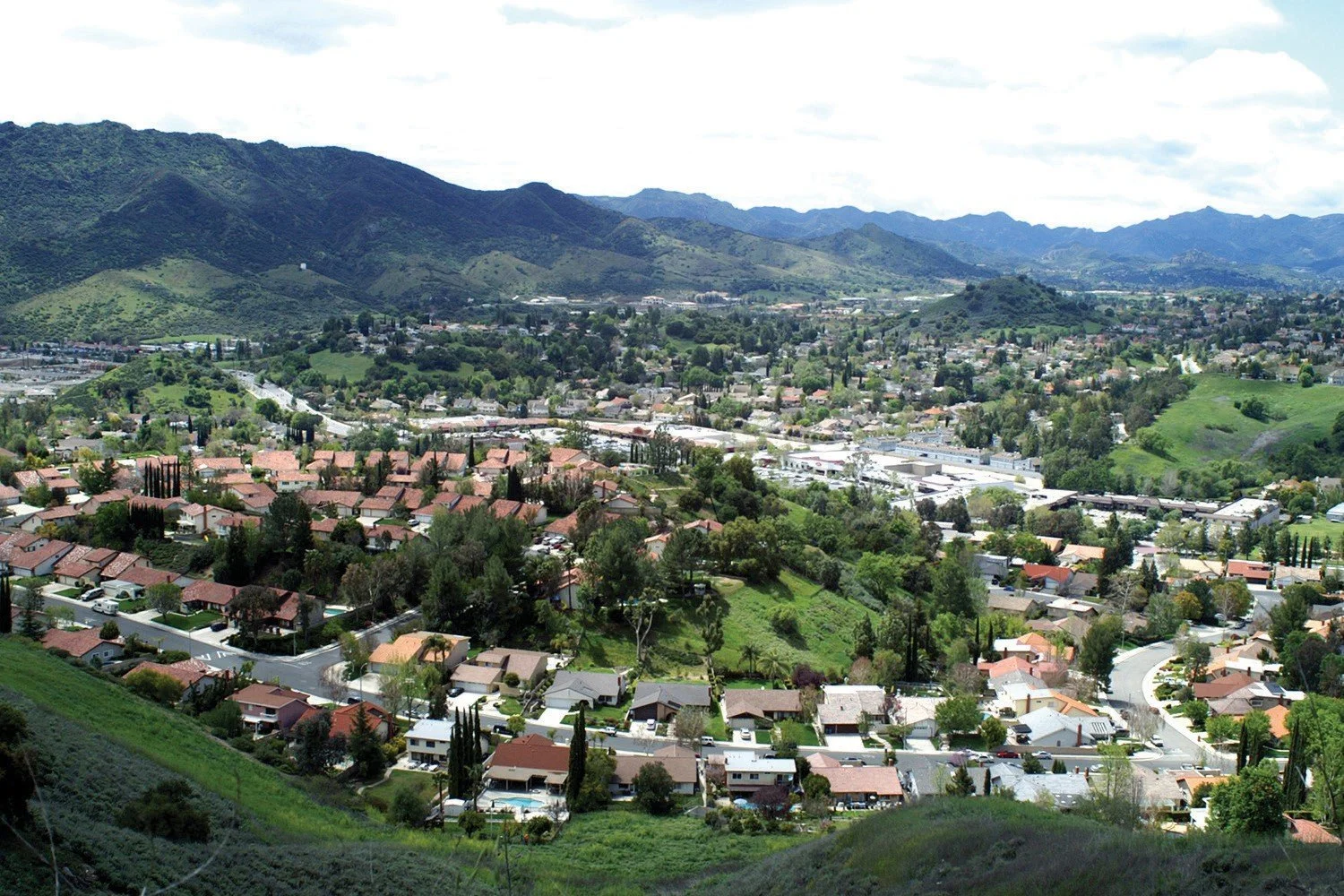 A view of Agoura Hills,CA  neighborhood nestled in a green valley with rolling hills and mountains in the background, featuring houses with red-tiled roofs, winding roads, and lush trees.