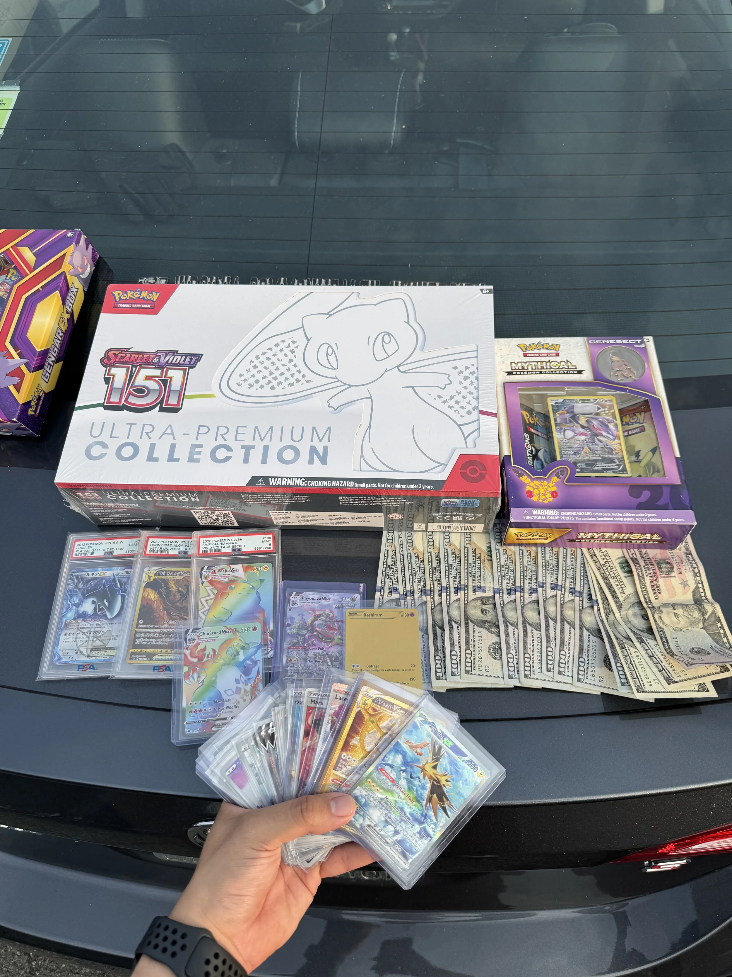 Collection of Pokémon trading cards, dollar bills, and Pokémon merchandise displayed on the trunk of a car, including a large box labeled 'Scarlet & Violet 151 Ultra-Premium Collection' and a smaller boxed set with Pokémon cards inside.