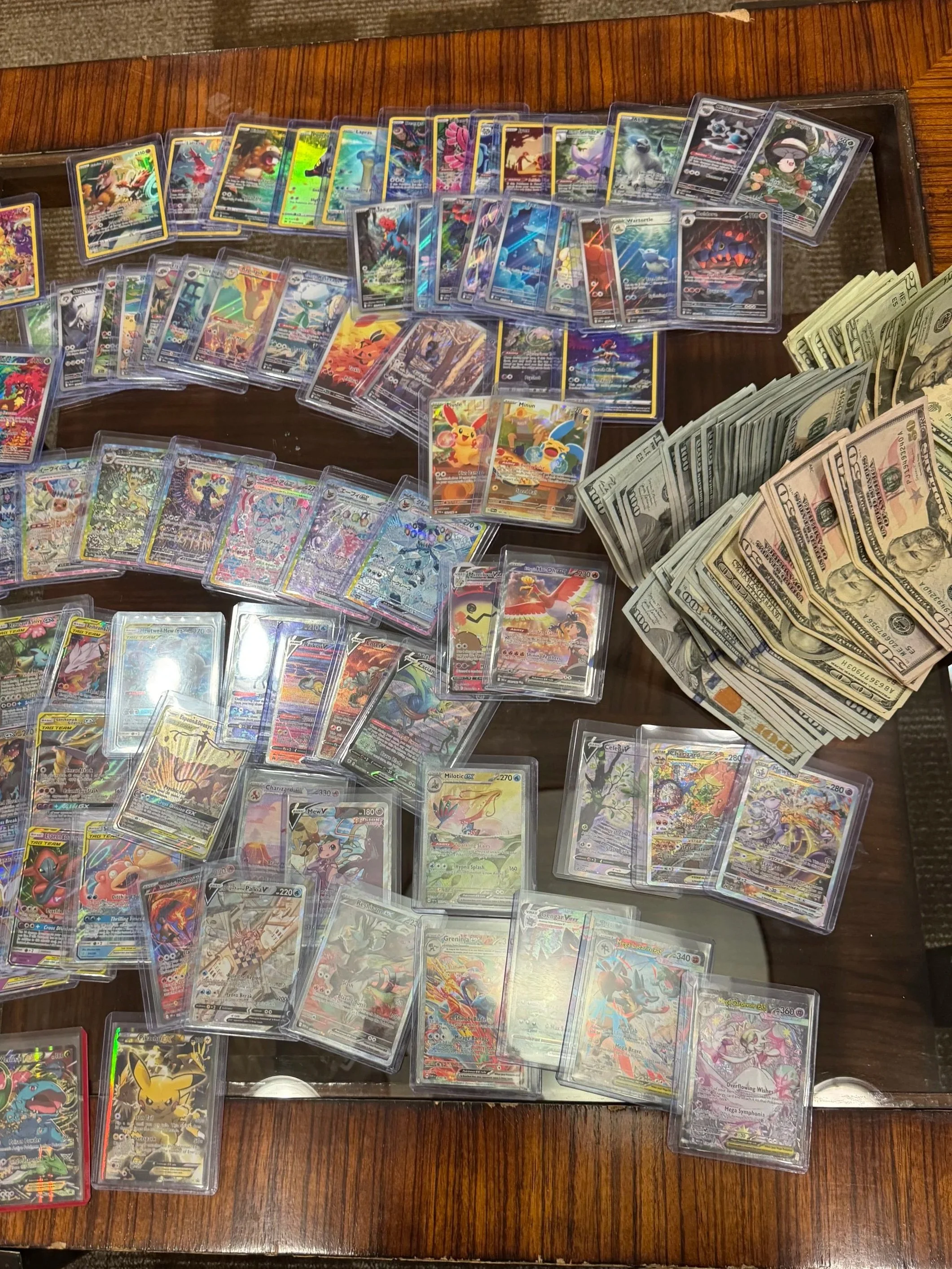 A large collection of Pokémon trading cards spread out on a wooden table alongside a pile of U.S. hundred-dollar bills.