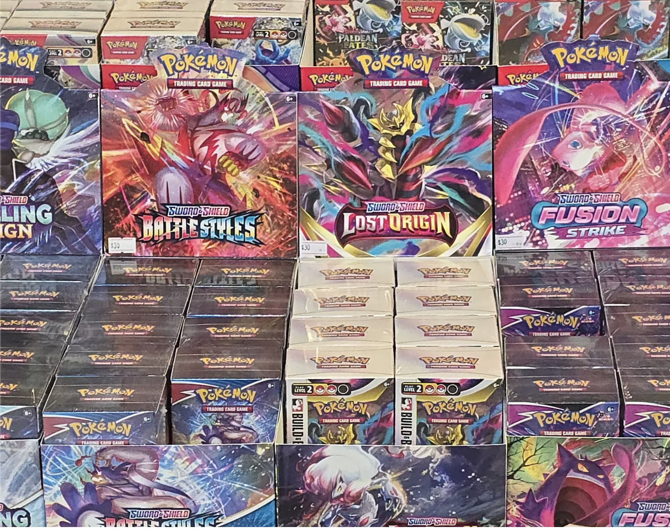 Display of Pokémon trading card game booster boxes featuring Sword and Shield sets, including Battle Styles, Lost Origin, Fusion Strike, and others, arranged on store shelves.