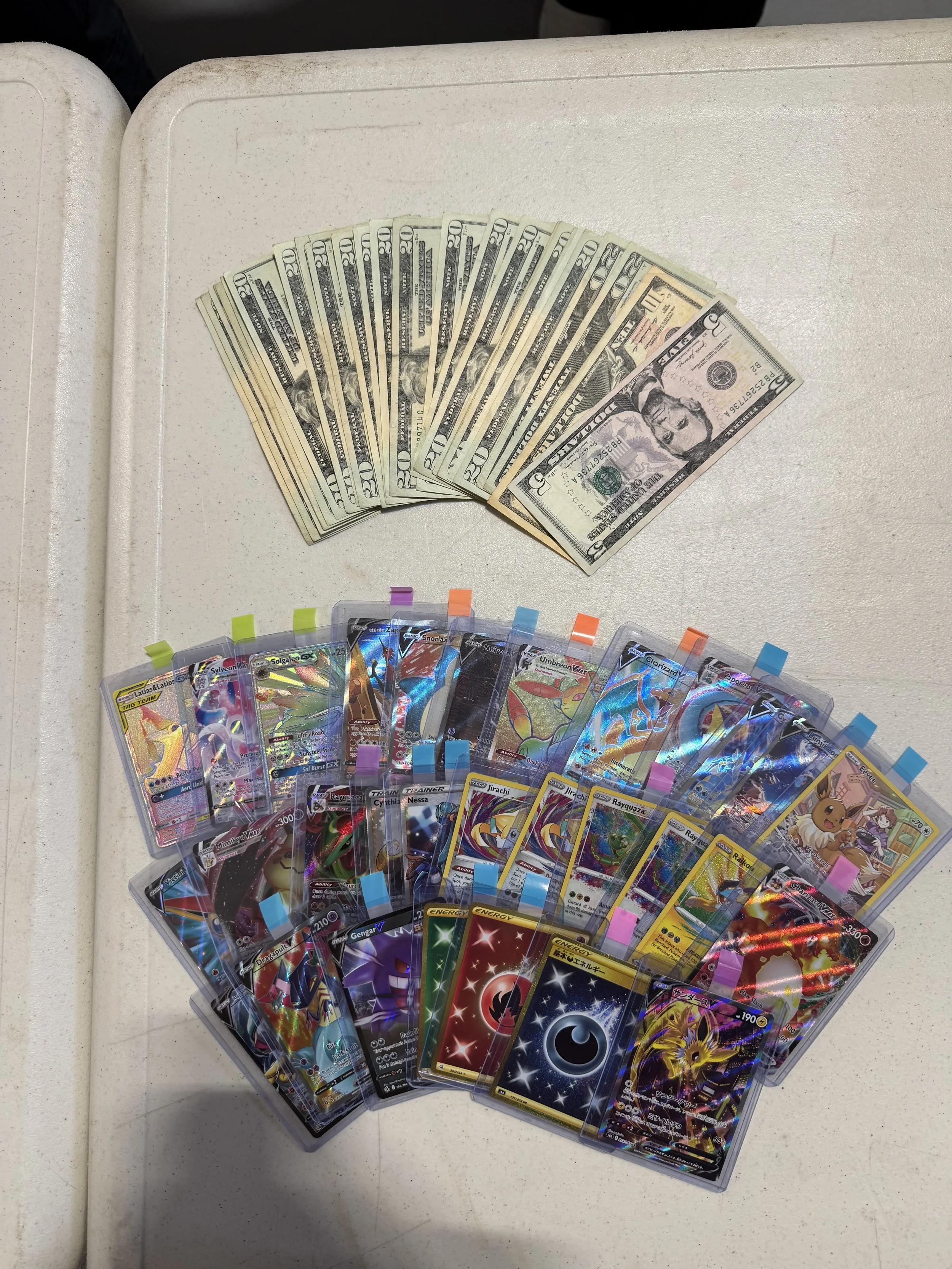A spread of U.S. dollar bills topped with a collection of Pokémon trading cards in plastic sleeves.