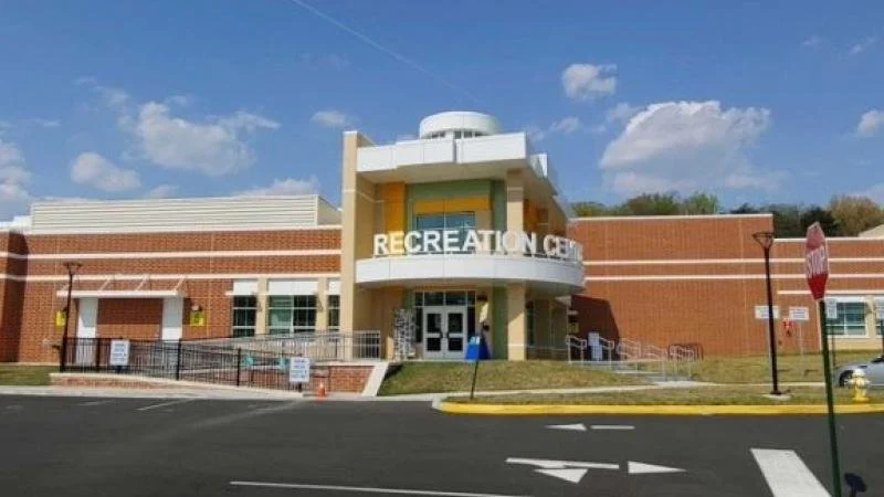 Front view of a recreation center building with a brick facade, glass doors, and a sign that says 'RECREATION CENTER' above the entrance. There are stairs and ramps for accessibility, a stop sign, and parking area in the foreground.