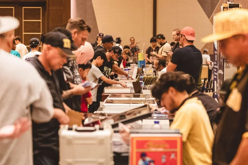 People browsing and purchasing items at a busy convention or trade show with tables displaying merchandise.