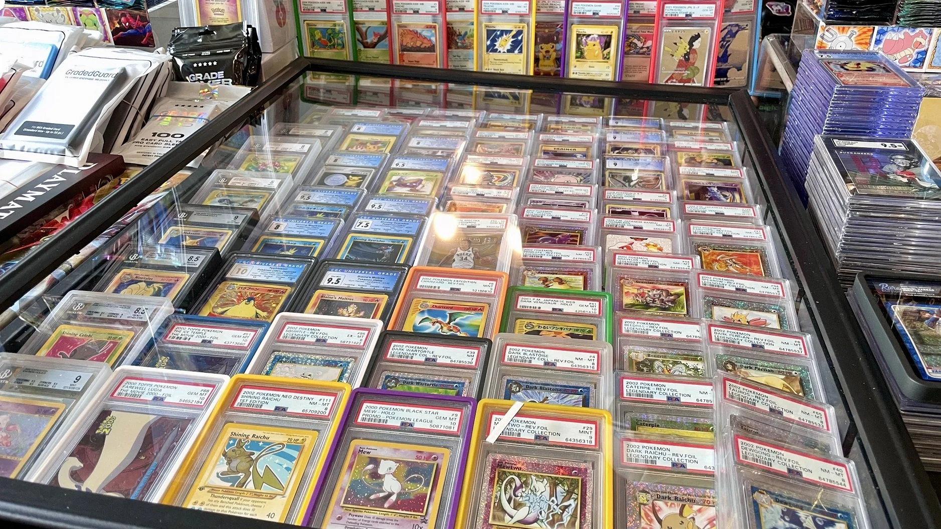 A display case filled with Pokémon trading cards at a store, with various cards in protective sleeves and some graded by professional grading companies, along with stacks of cards outside the case.