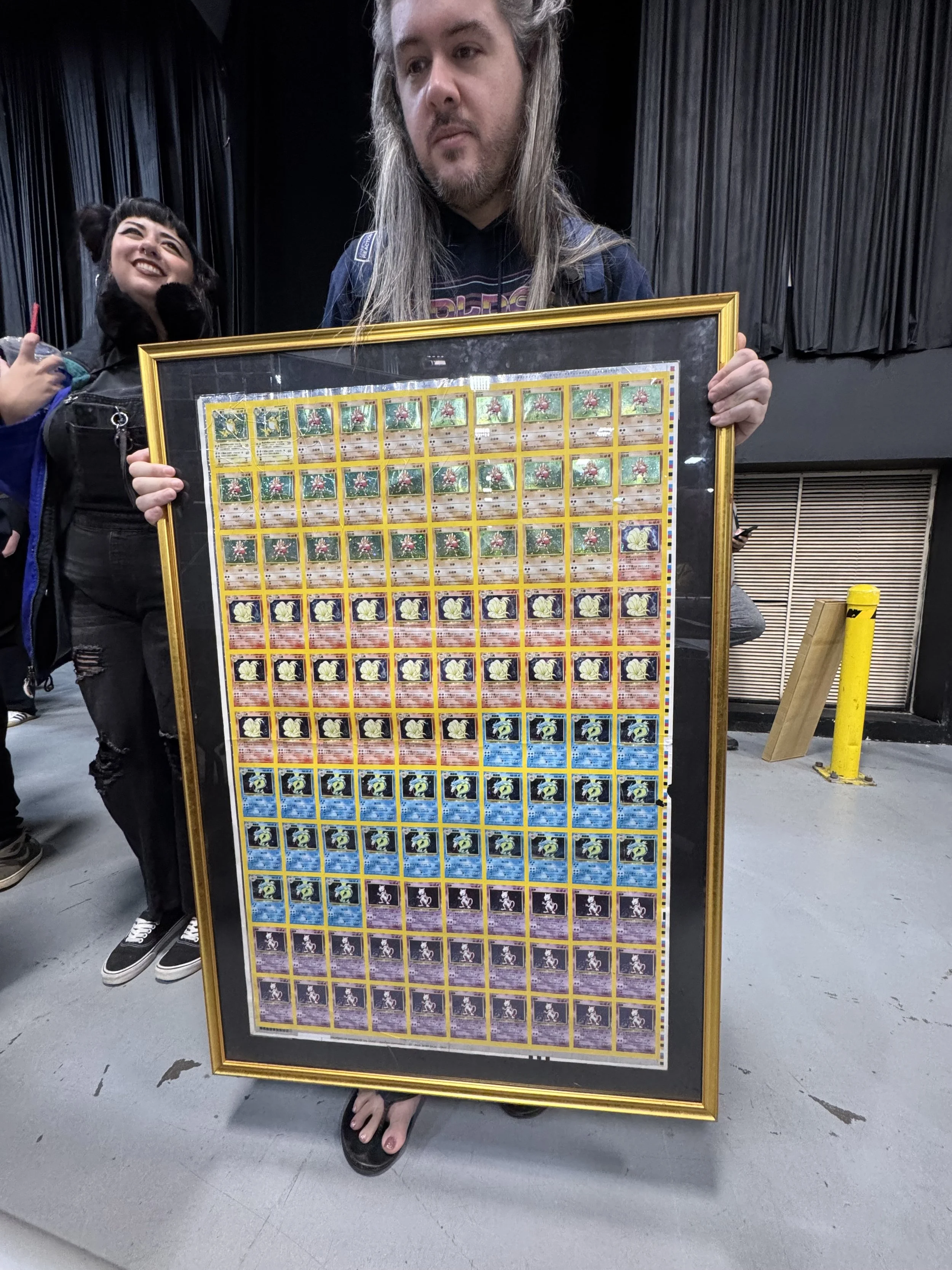 Person with long hair holding a framed display of Pokémon trading cards, with a woman standing nearby, smiling, in an indoor setting.