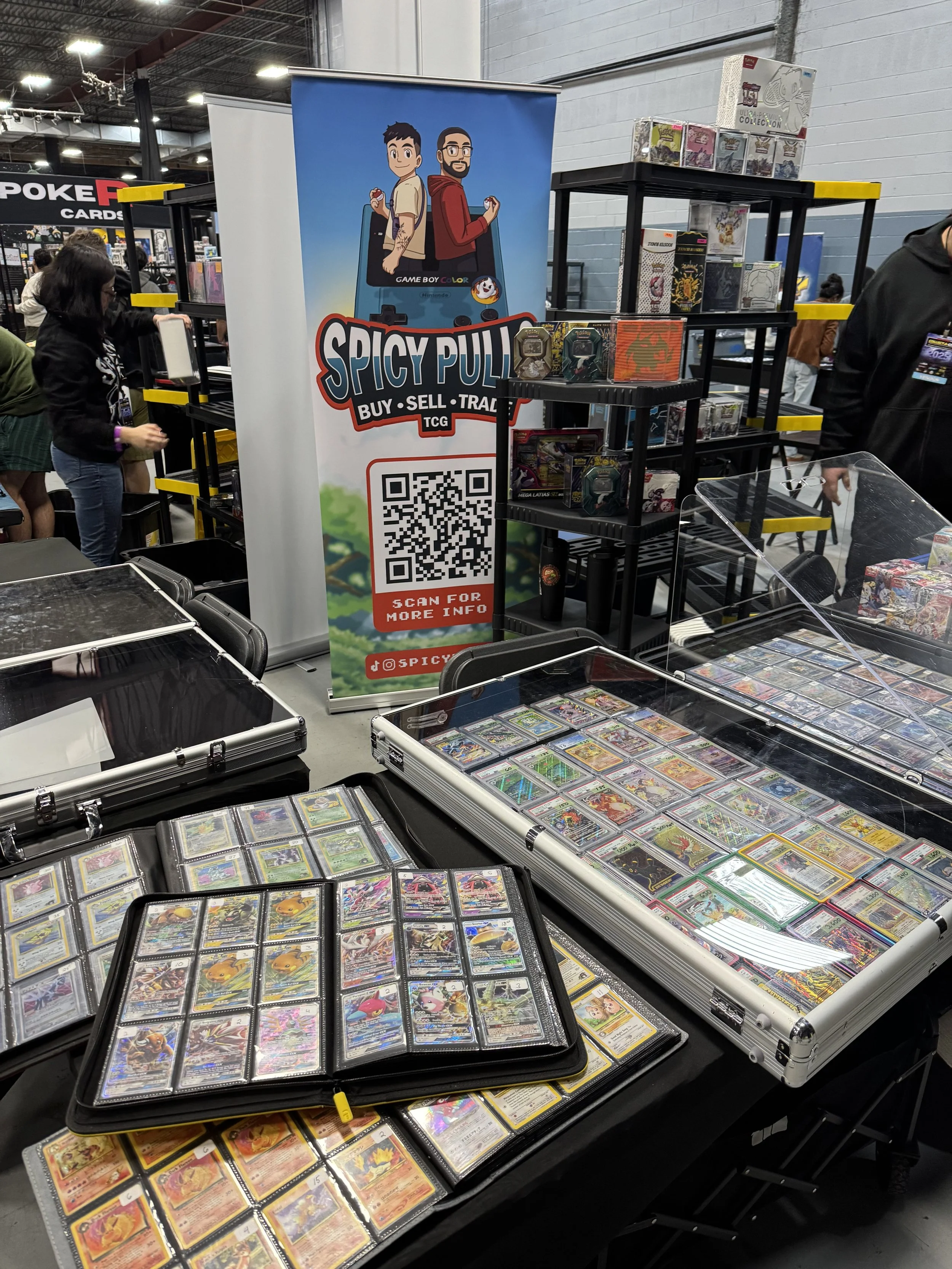 Table displays various Pokémon trading cards in binder pages and cases, with a logo banner for Spicy Pull, a company for buying, selling, and trading cards, featuring cartoon characters at a convention or expo.